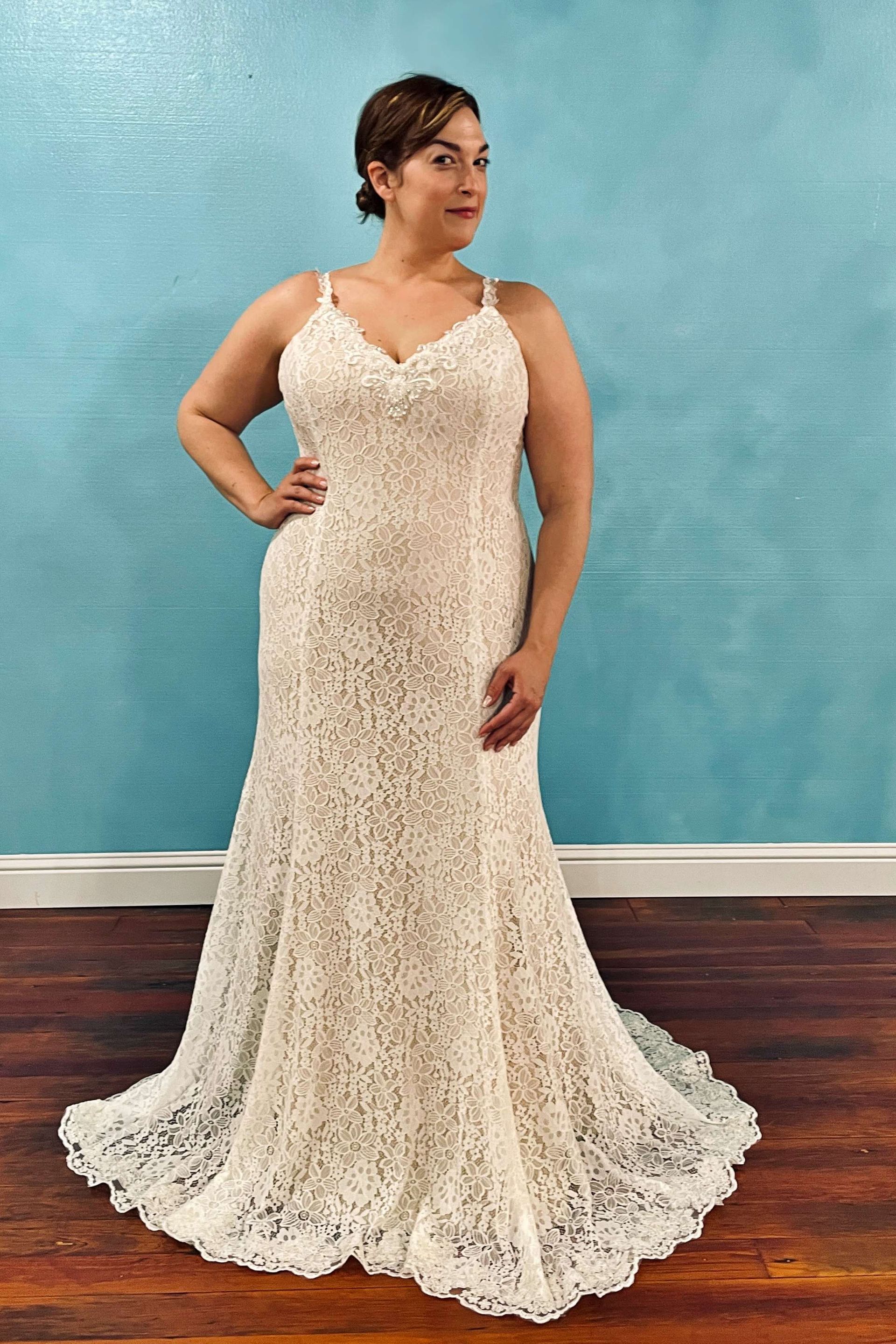 Woman in a white lace wedding dress, poses with hand on hip. Against a turquoise wall, on a wood floor.