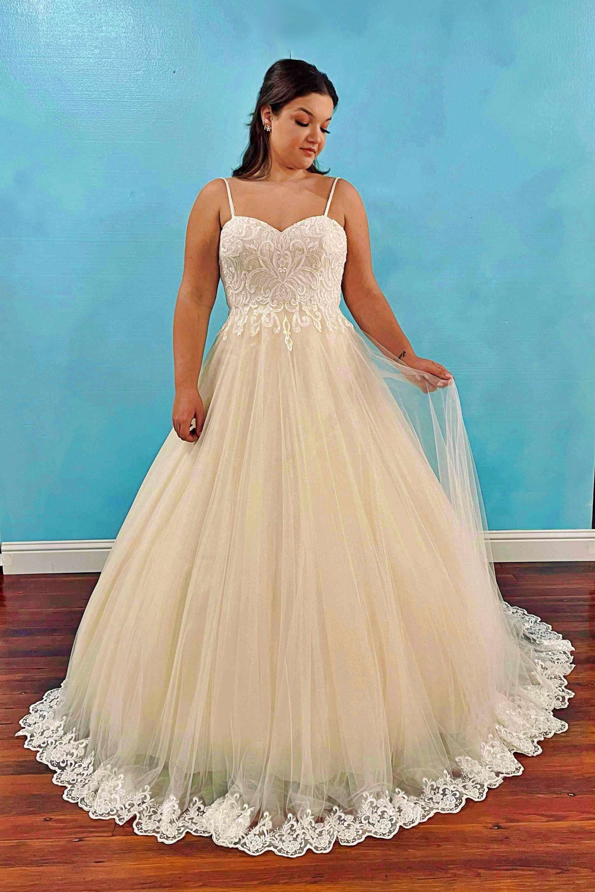 Woman in a cream-colored wedding dress with spaghetti straps, lace bodice, and tulle skirt, smiling, indoor setting.