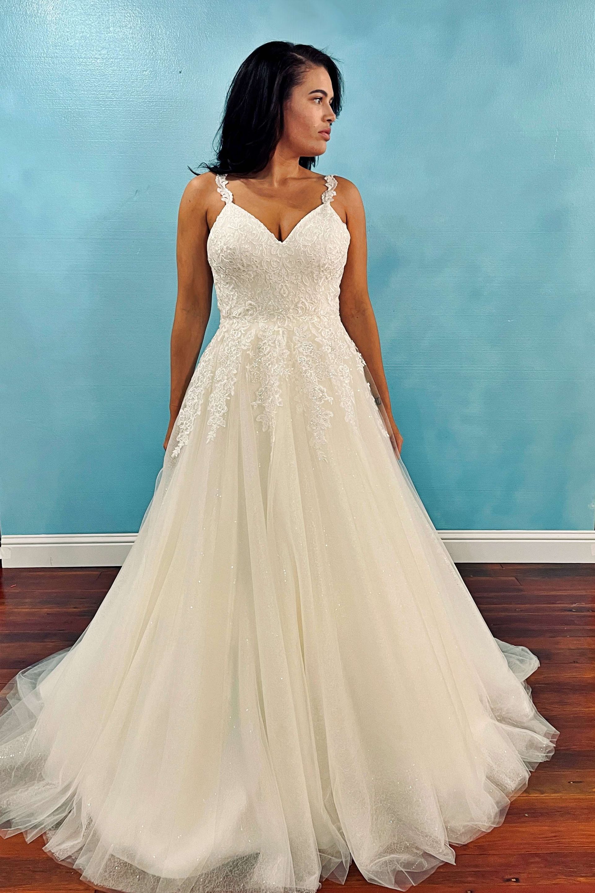 Woman in a white wedding dress with beaded bodice, spaghetti straps, and a full skirt, standing.