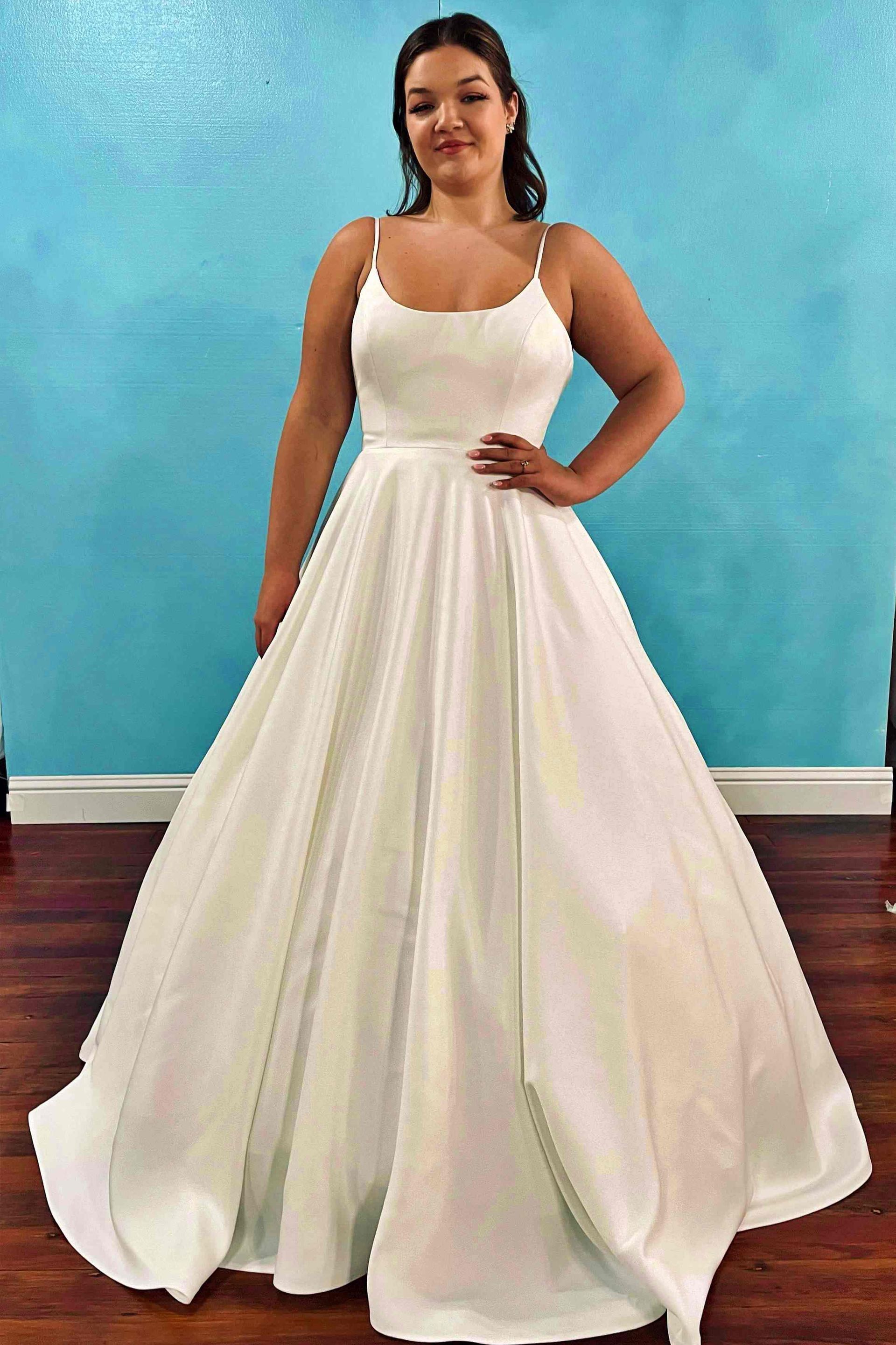 Woman in a white wedding dress, posing with hand on hip. Dress has thin straps and a full skirt. Blue wall.