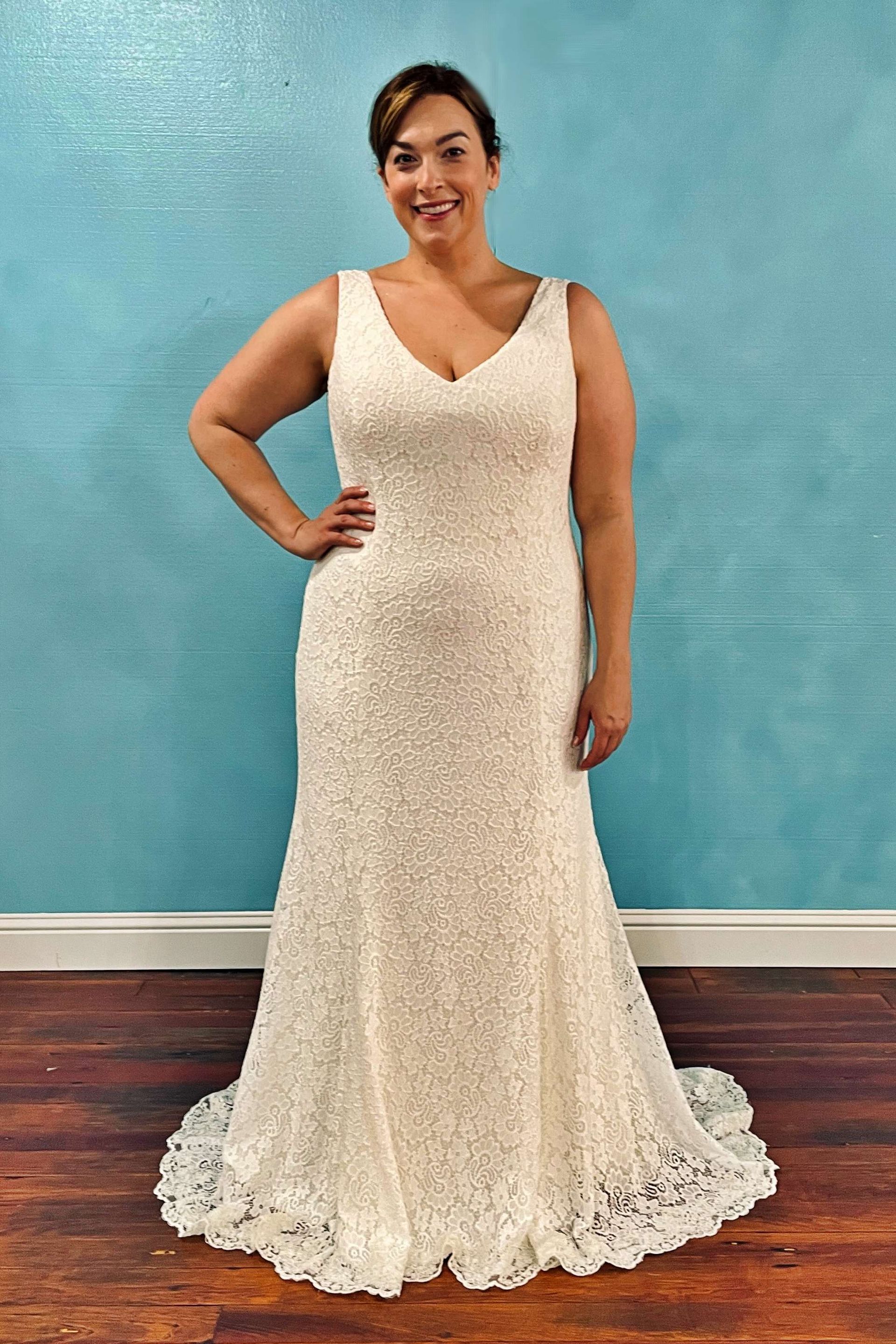Woman in a white lace wedding dress with a V-neck, posing with hand on hip in front of a blue wall.