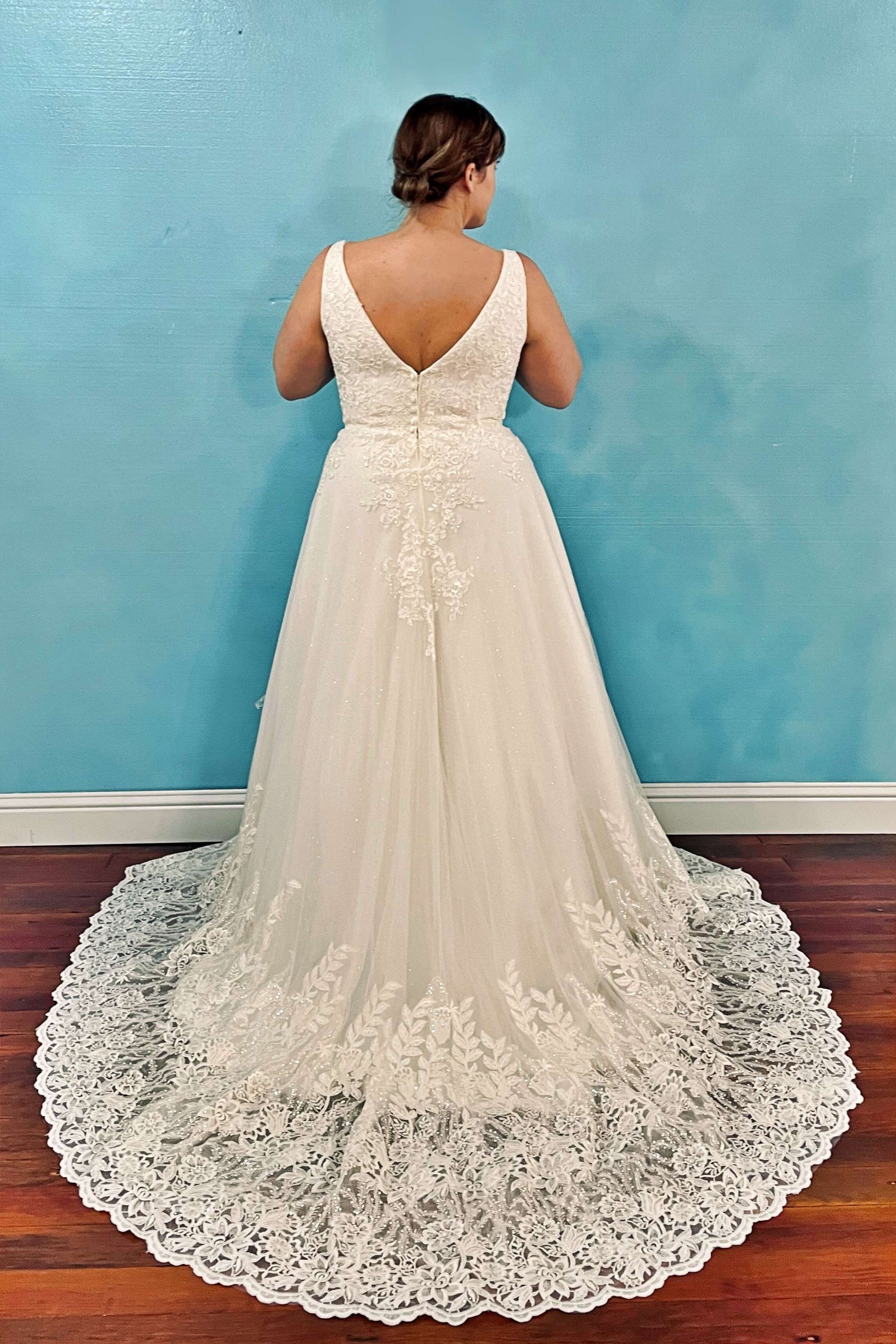 Woman in a white wedding dress with lace detail, standing in front of a blue wall, looking away.