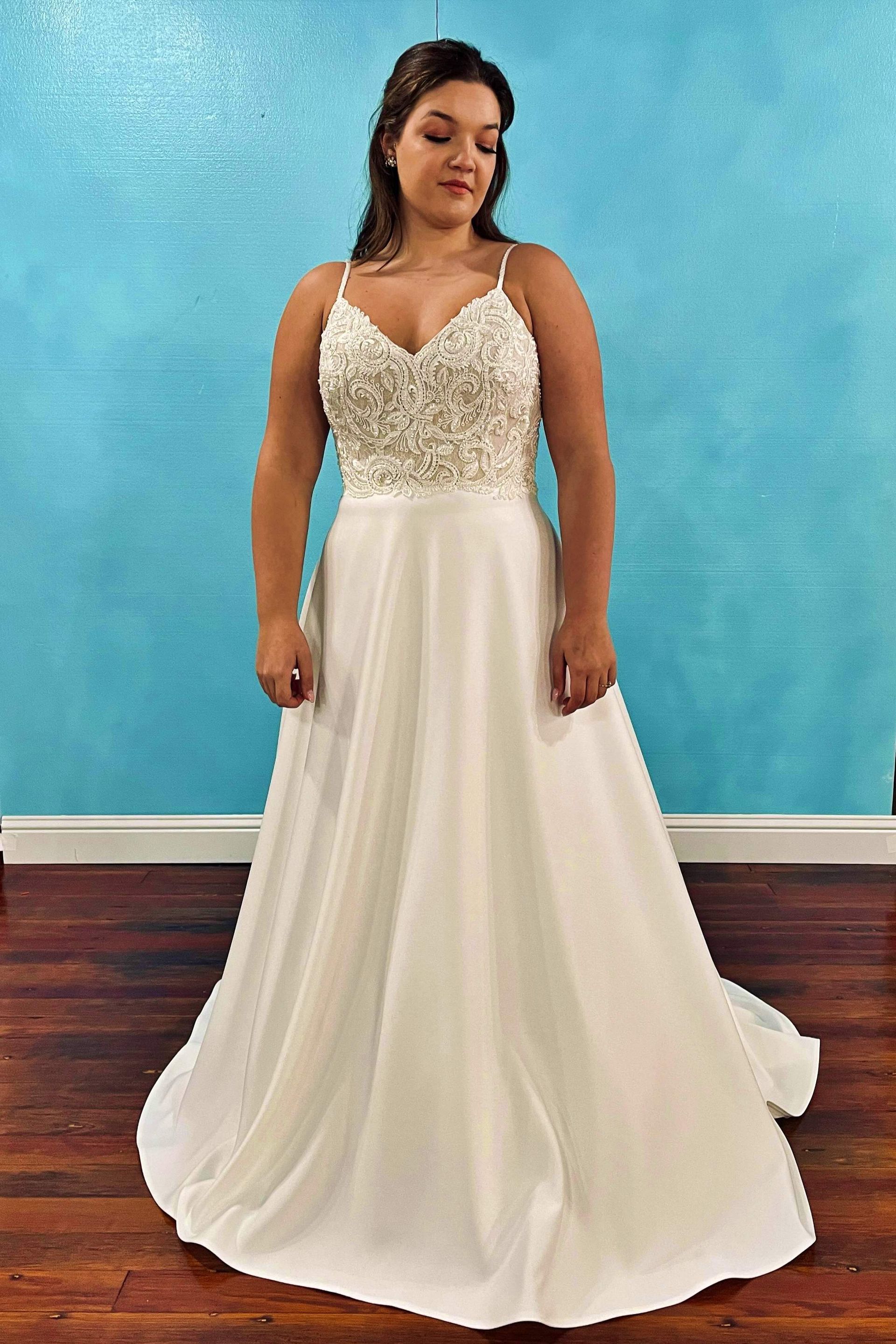 Woman wearing a white wedding dress with a beaded bodice and flowing skirt, standing in front of a blue wall.