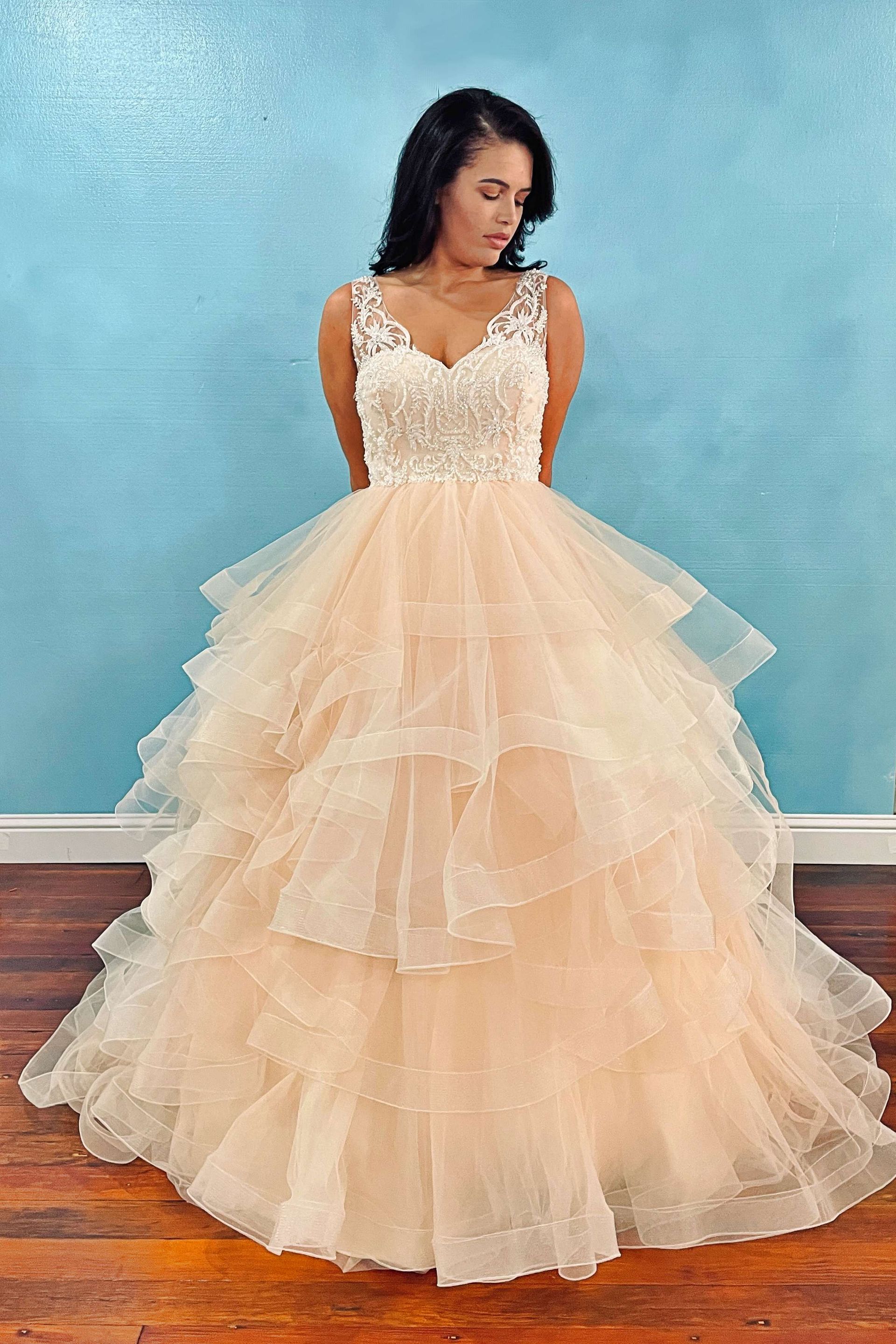Woman in a peach-colored, layered ballgown, with a beaded bodice, standing in front of a blue wall.