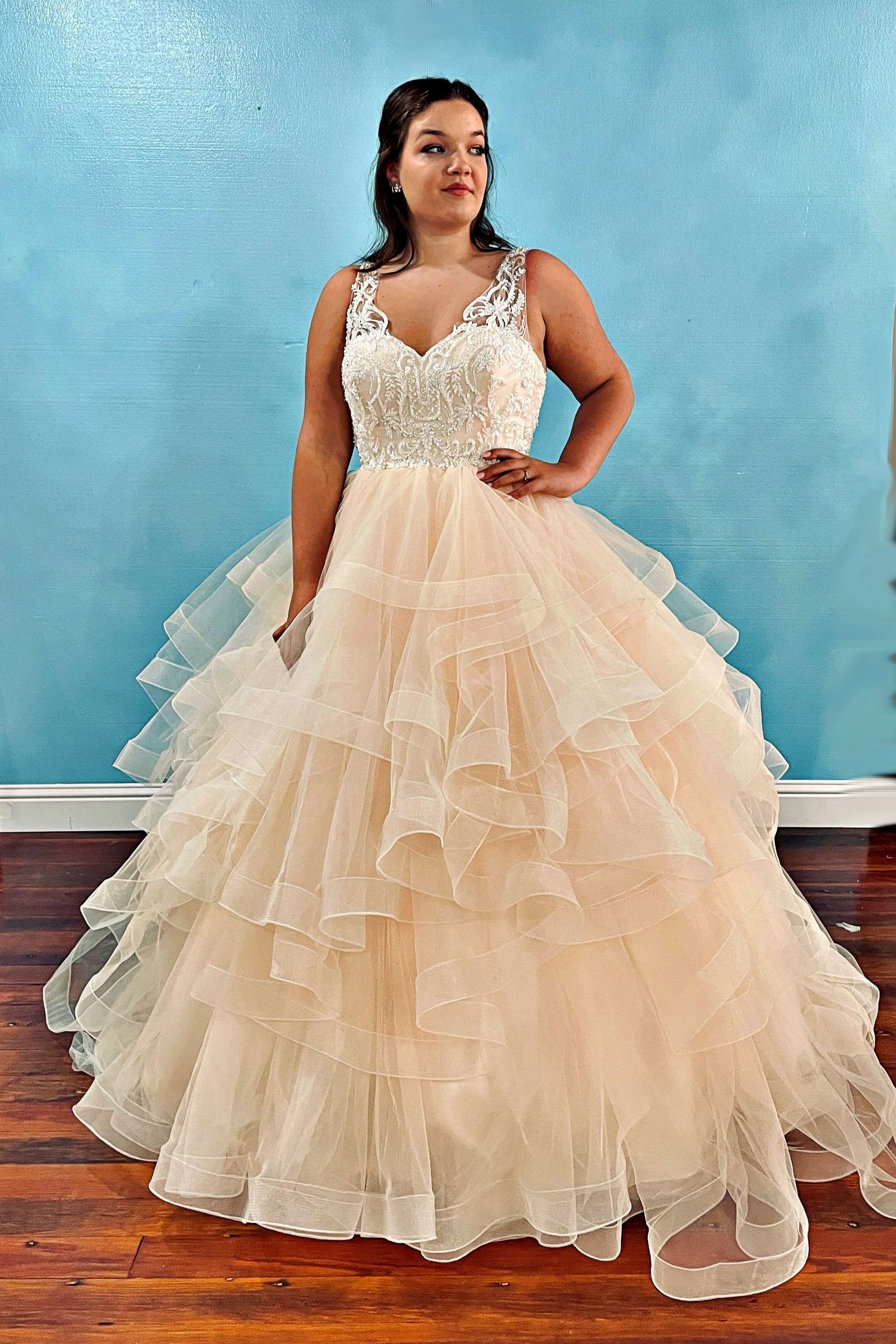 Woman in a formal, layered peach gown poses against a light blue wall.