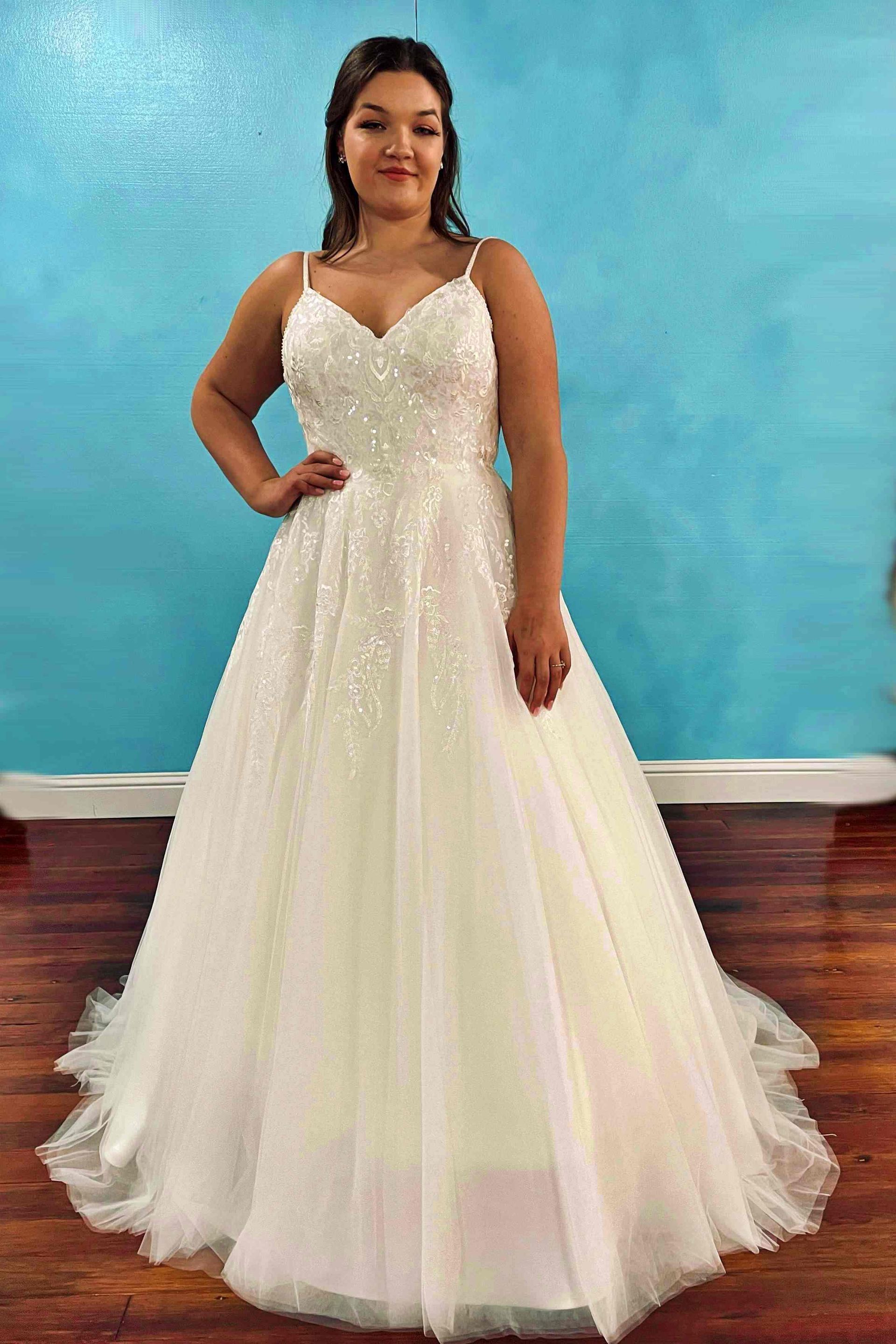Woman in a white wedding dress with spaghetti straps and a sparkly bodice, standing in front of a blue background.