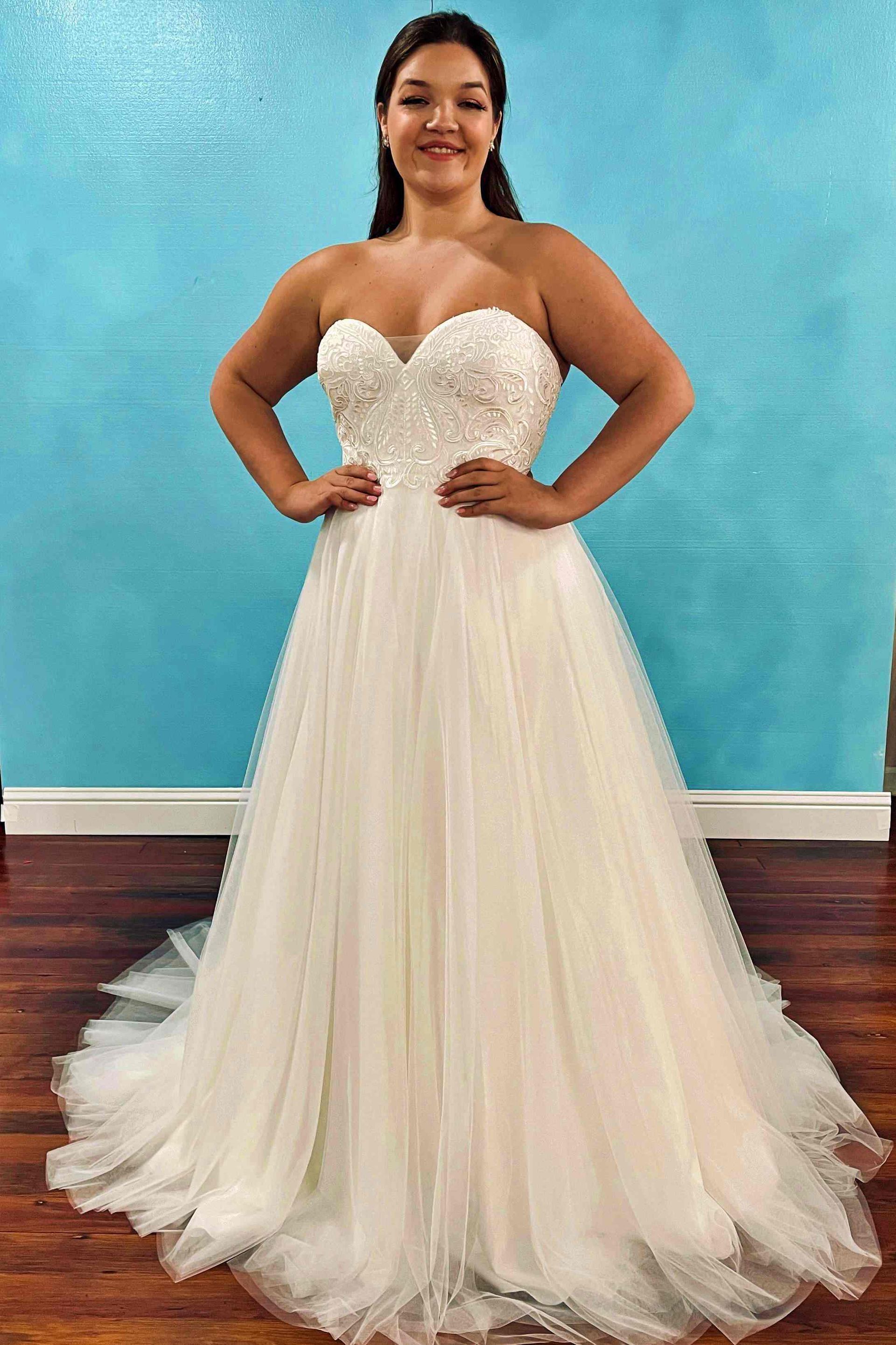 Woman in a white strapless wedding dress, hands on hips, standing in front of a blue wall.