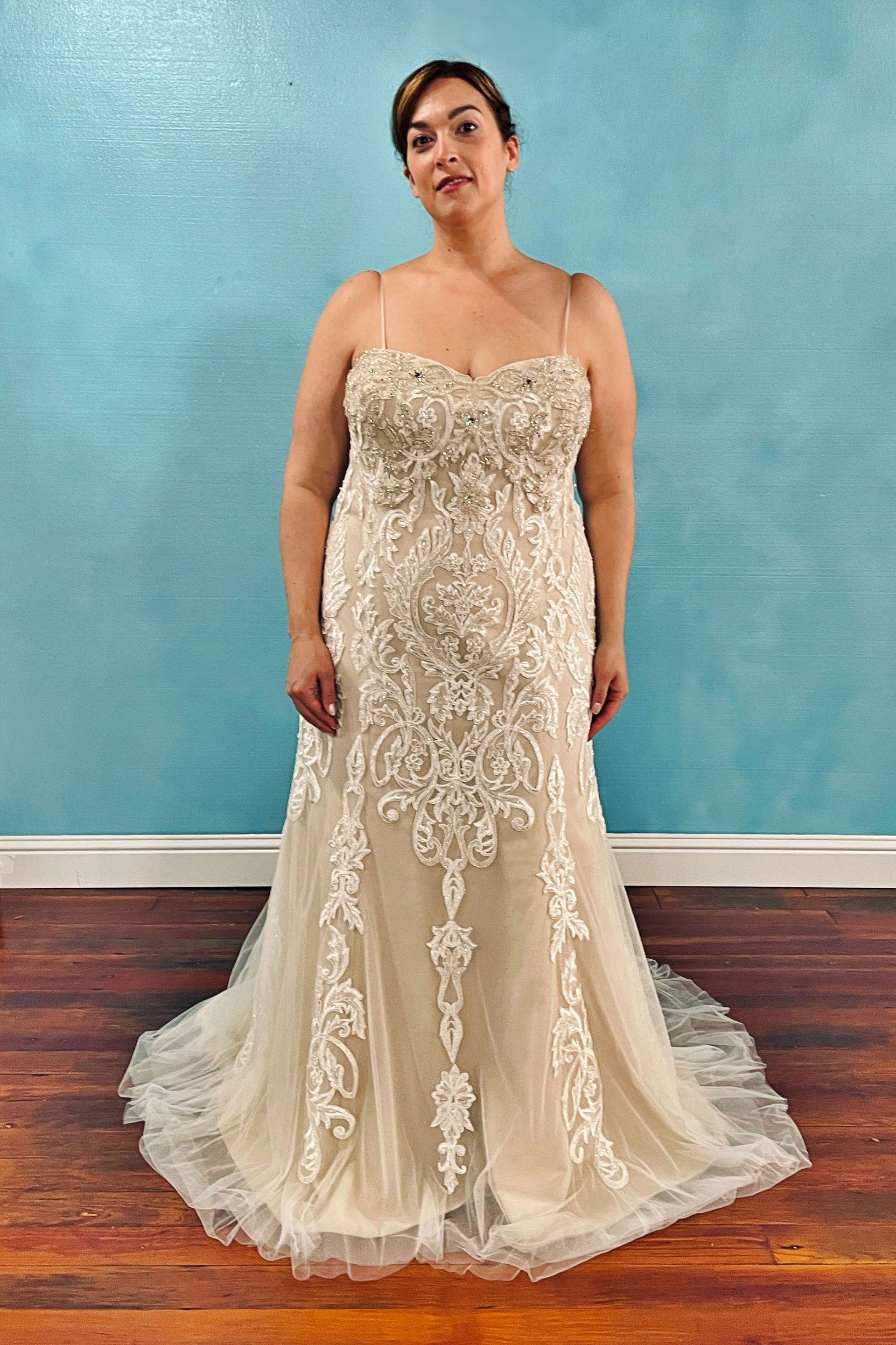 Woman in a wedding gown stands against a blue wall. The dress is ivory with lace detailing and a flowing skirt.