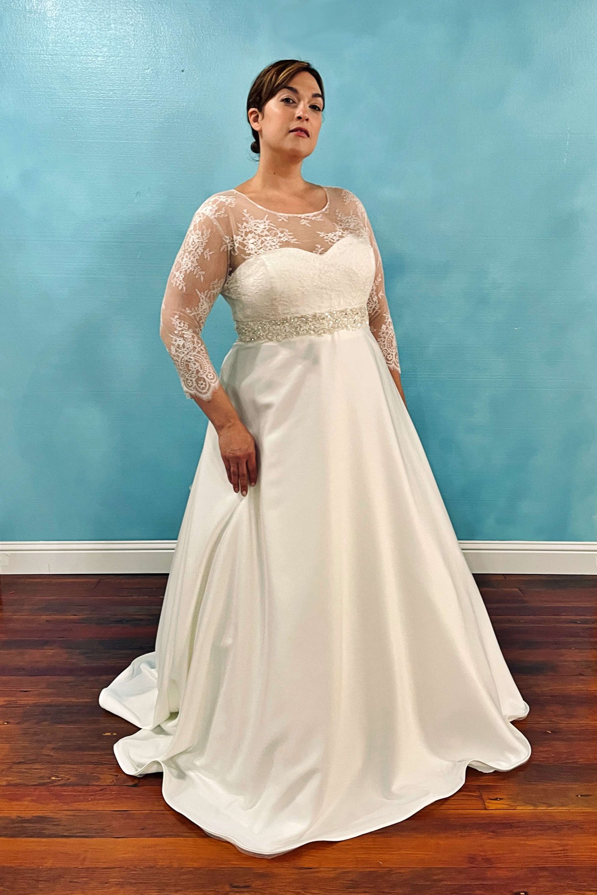 Woman in a white wedding dress with lace sleeves and a beaded belt, standing against a blue wall.