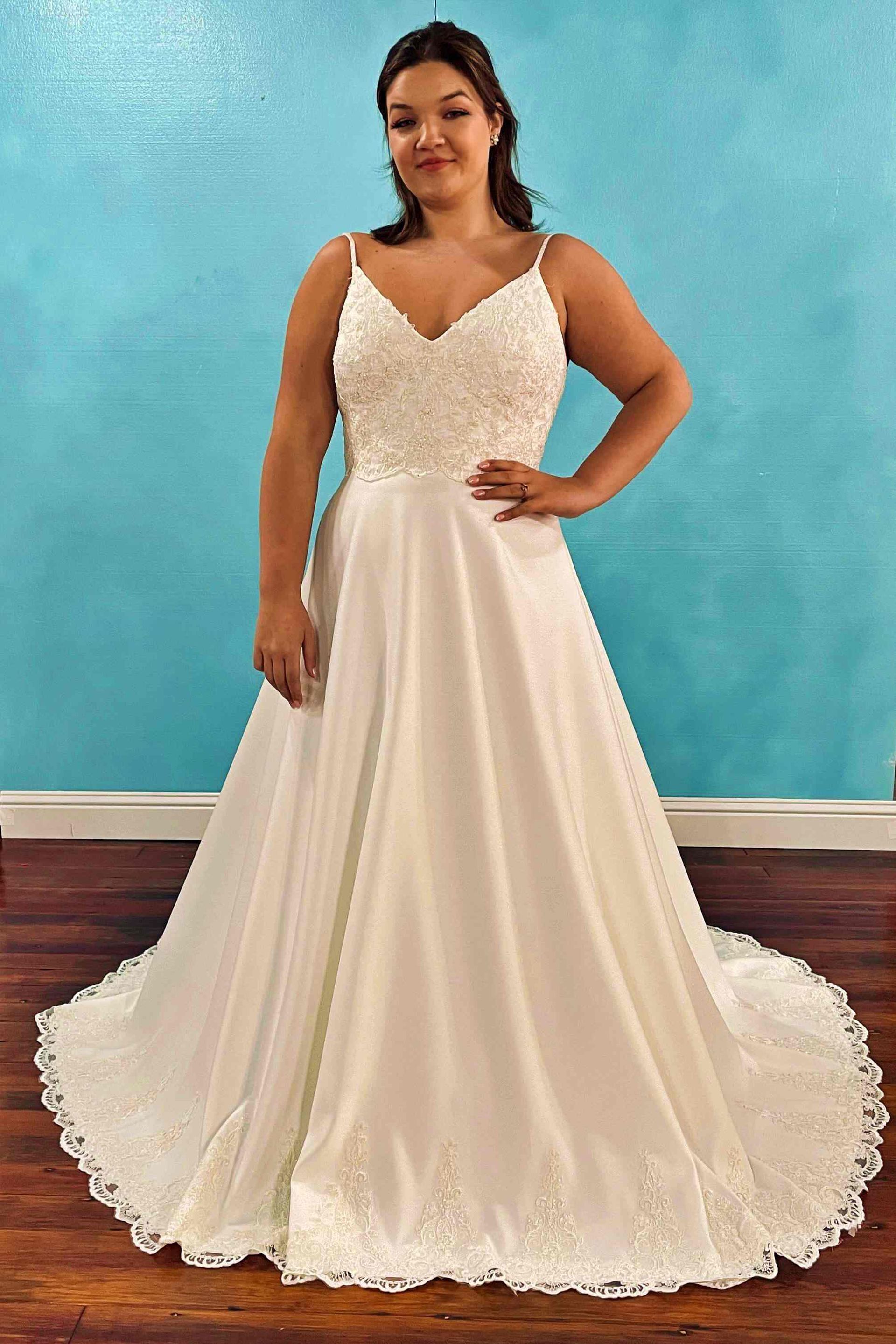 Woman wearing a white wedding dress with beaded bodice, spaghetti straps, and a-line skirt; standing in front of a blue wall.