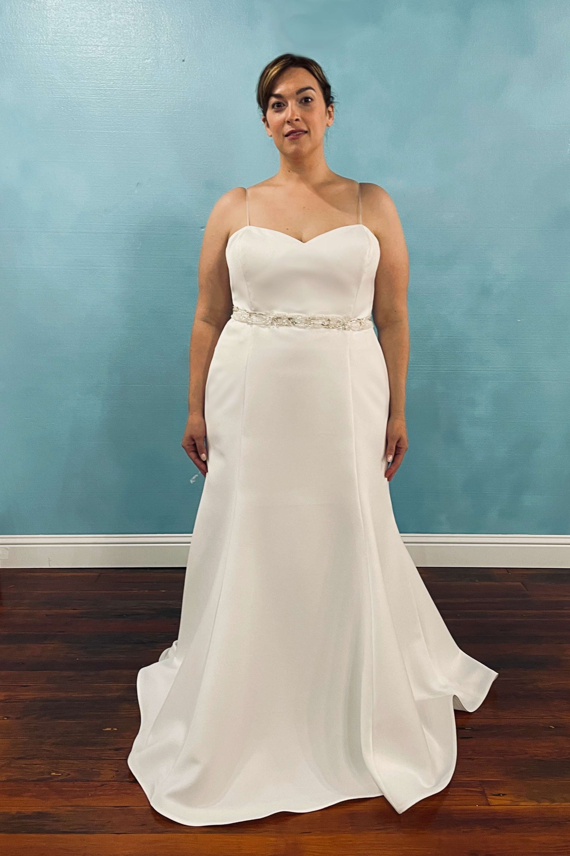 Woman in a white wedding gown with a beaded belt and thin straps, standing in front of a blue wall.