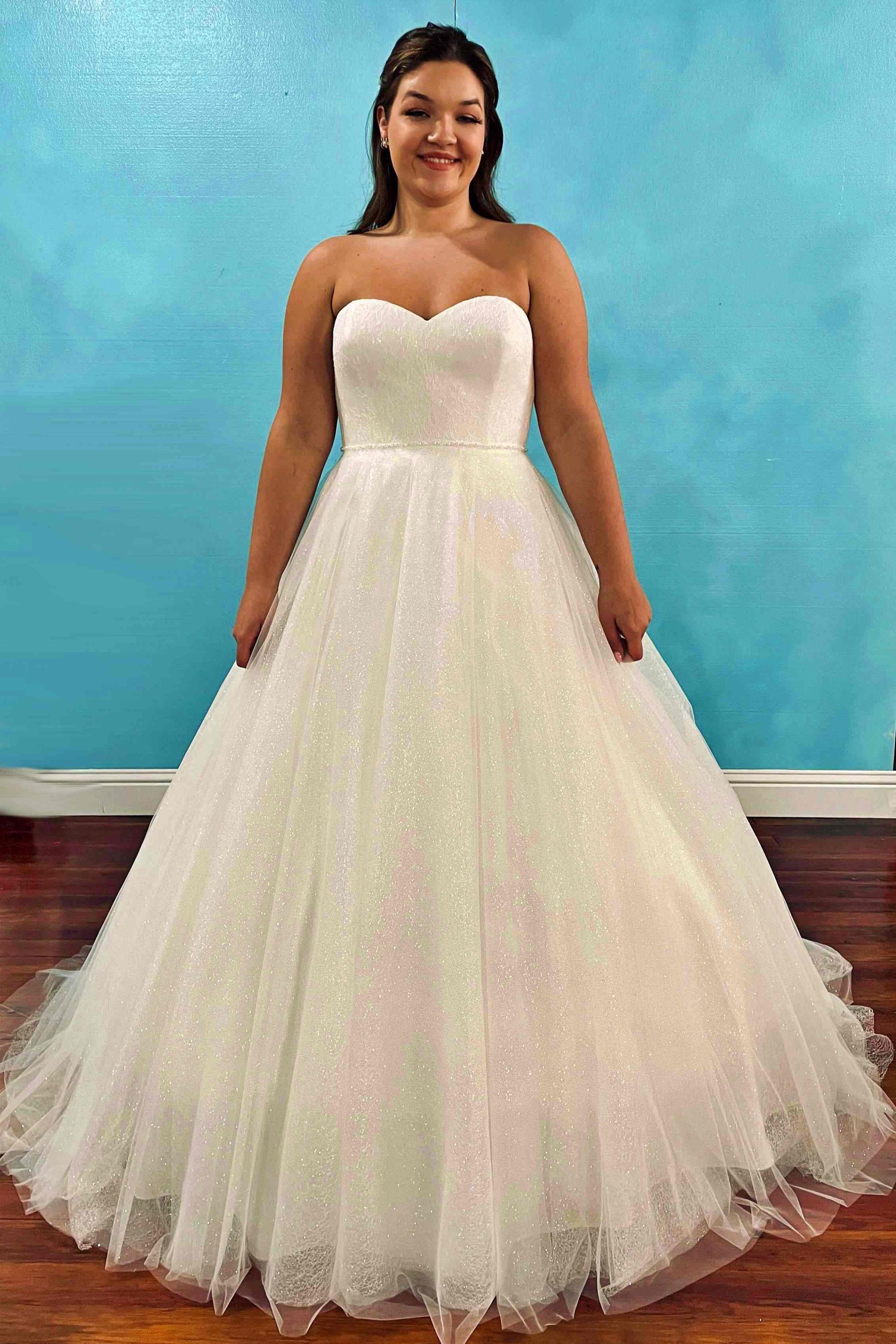 Woman in strapless white sparkly ballgown smiling, standing in front of blue wall and wood floor.