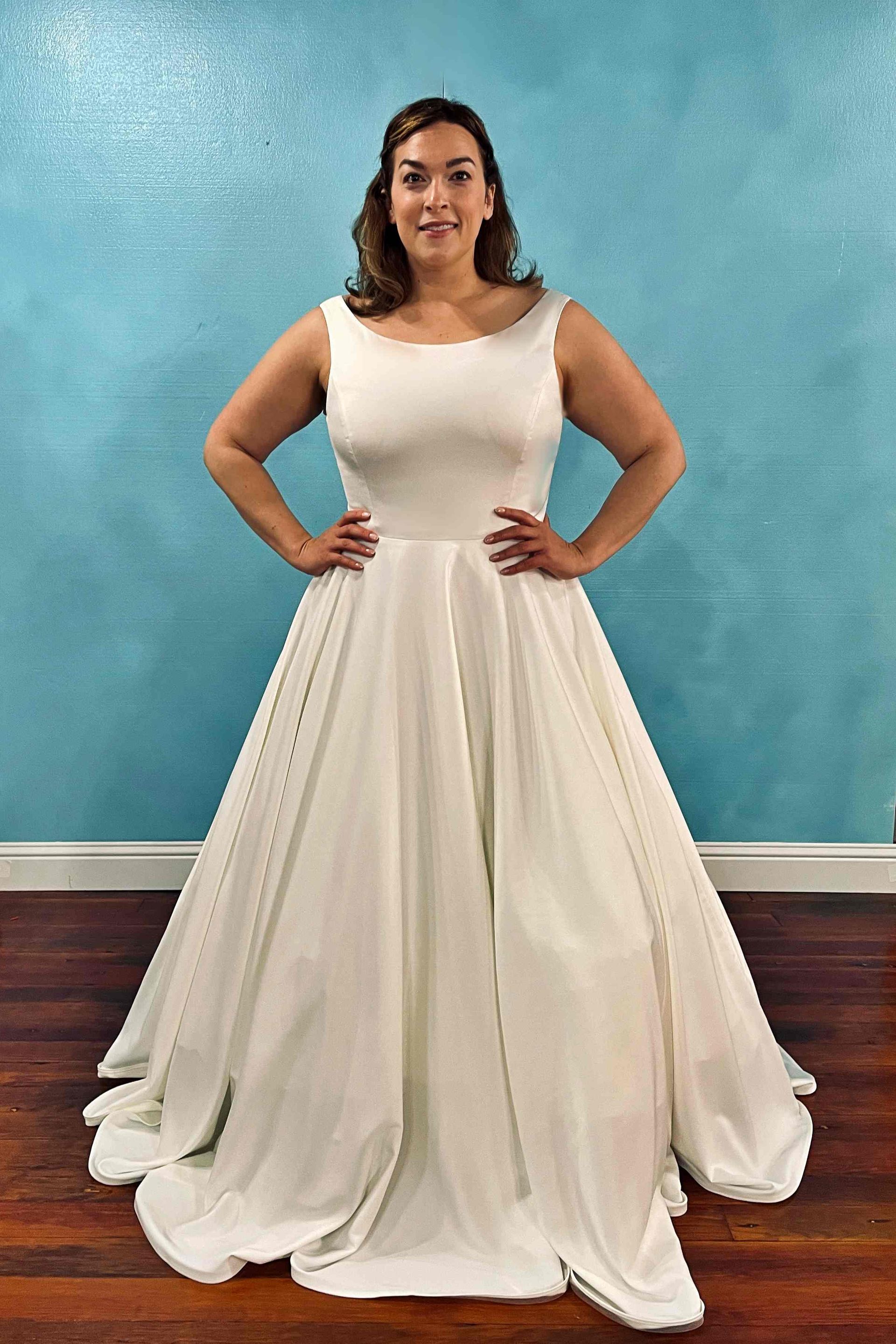 Woman in white wedding dress with hands on hips, standing in front of a blue wall.
