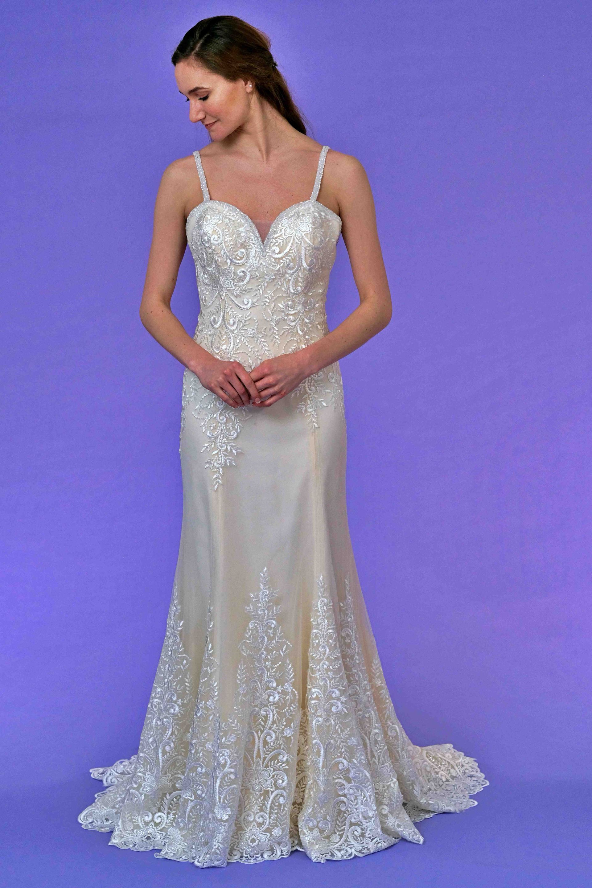 Woman wearing a white, beaded wedding dress with spaghetti straps and a mermaid skirt, smiling, on purple background.