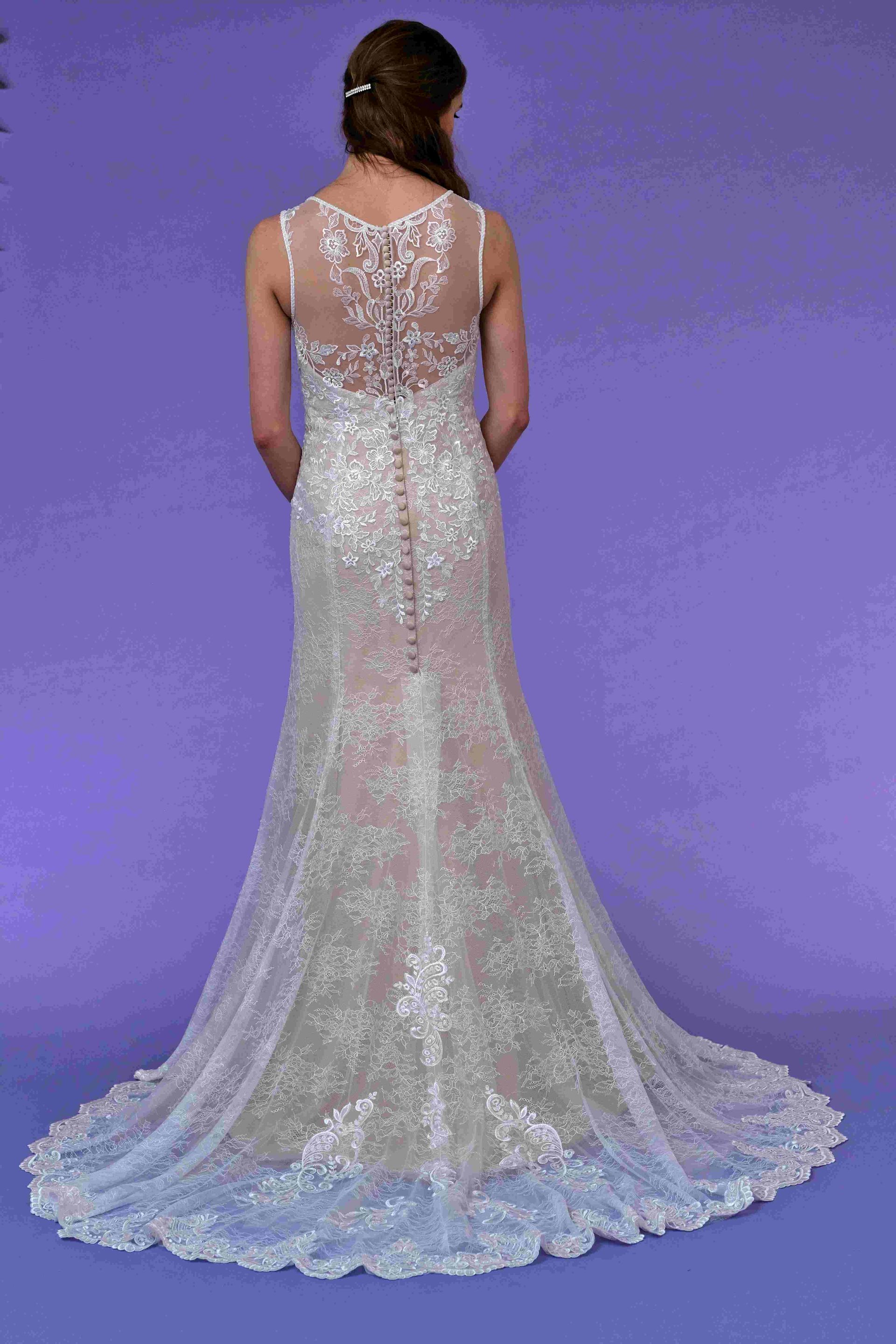 Back view of a bride in a sparkling, detailed white lace wedding gown with a train, standing against a purple backdrop.