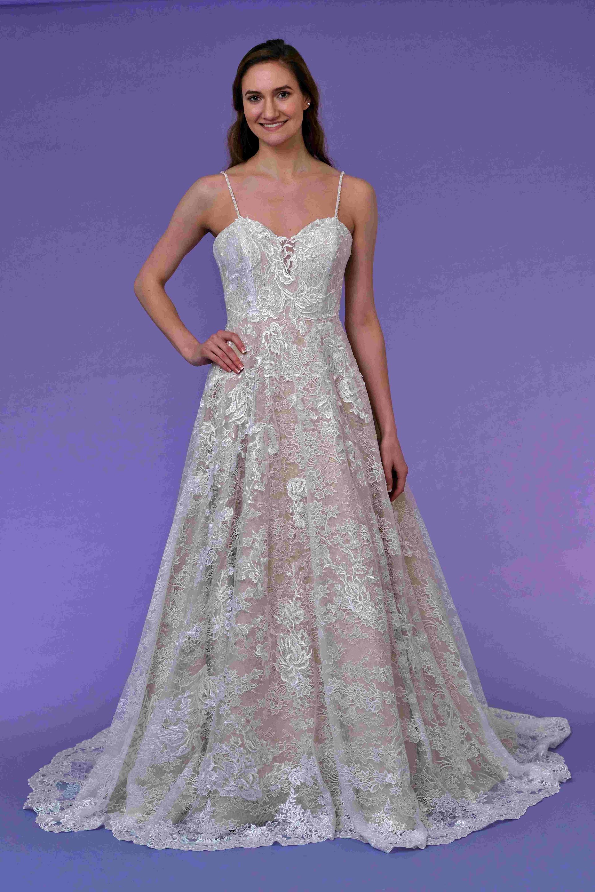 Woman wearing a white, lacy wedding gown with spaghetti straps, standing in front of a purple backdrop.