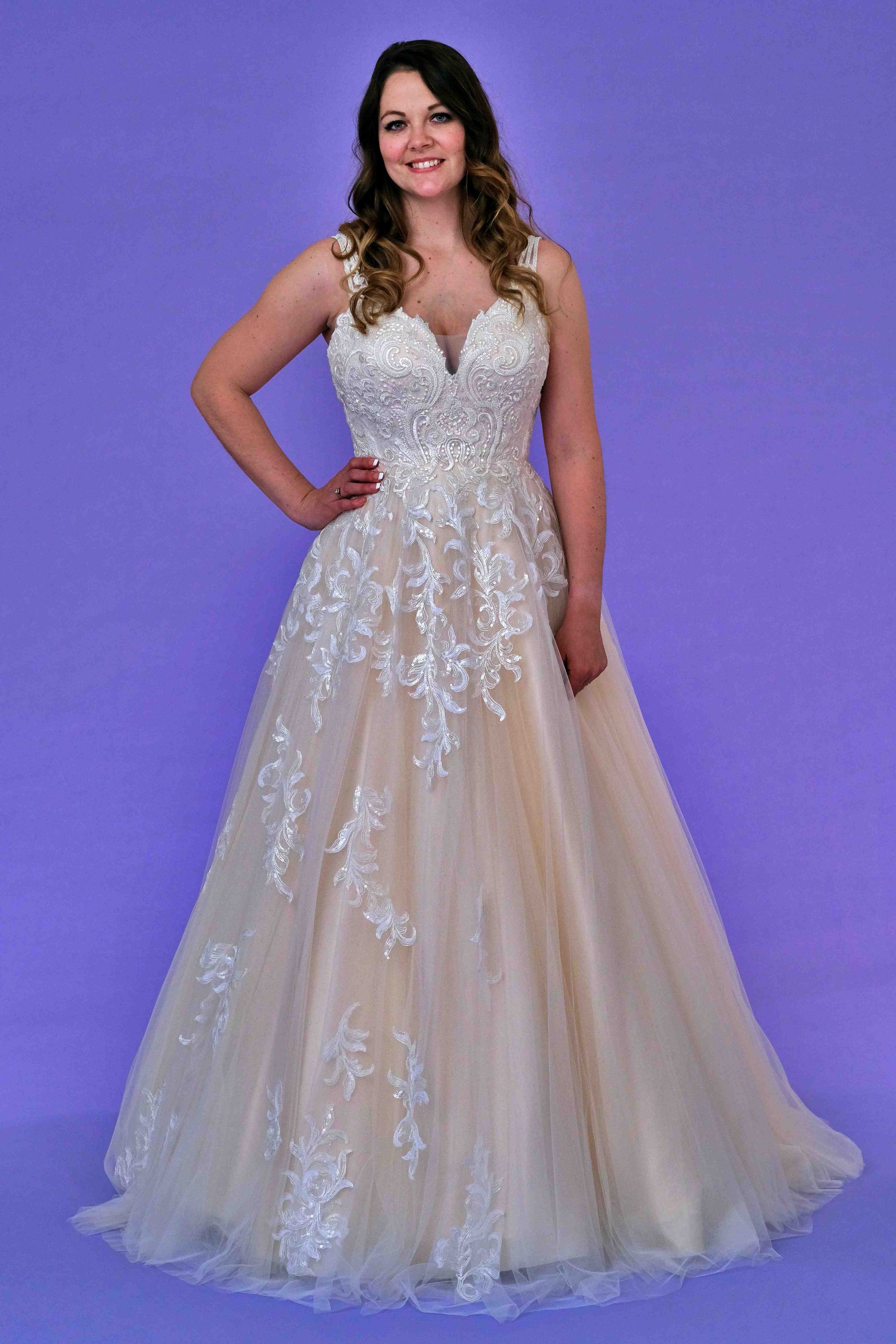 Woman in a champagne-colored wedding dress with white lace detailing, standing on a purple background, smiling.