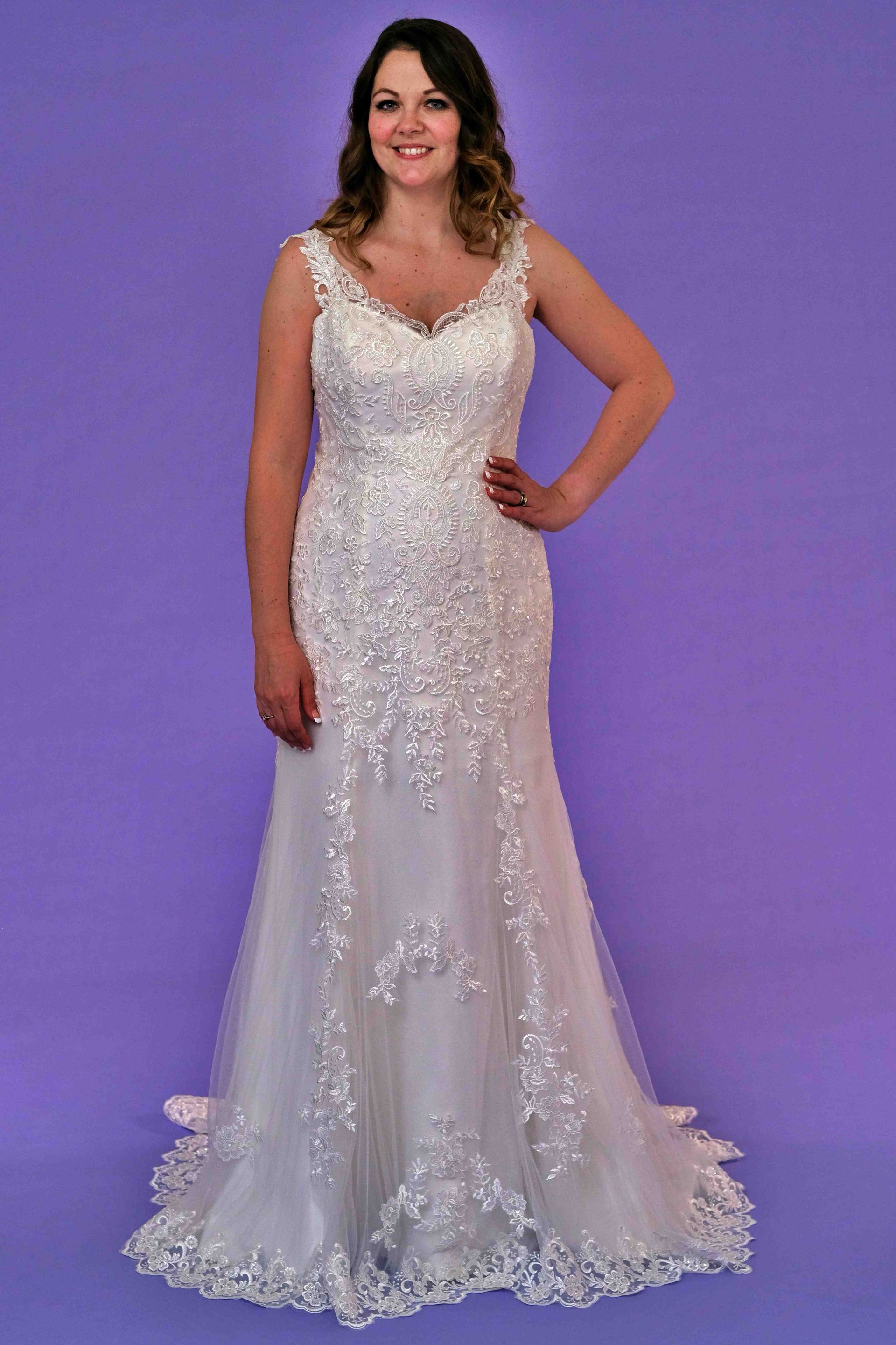 Woman in a white beaded wedding dress with lace details, standing against a purple background.
