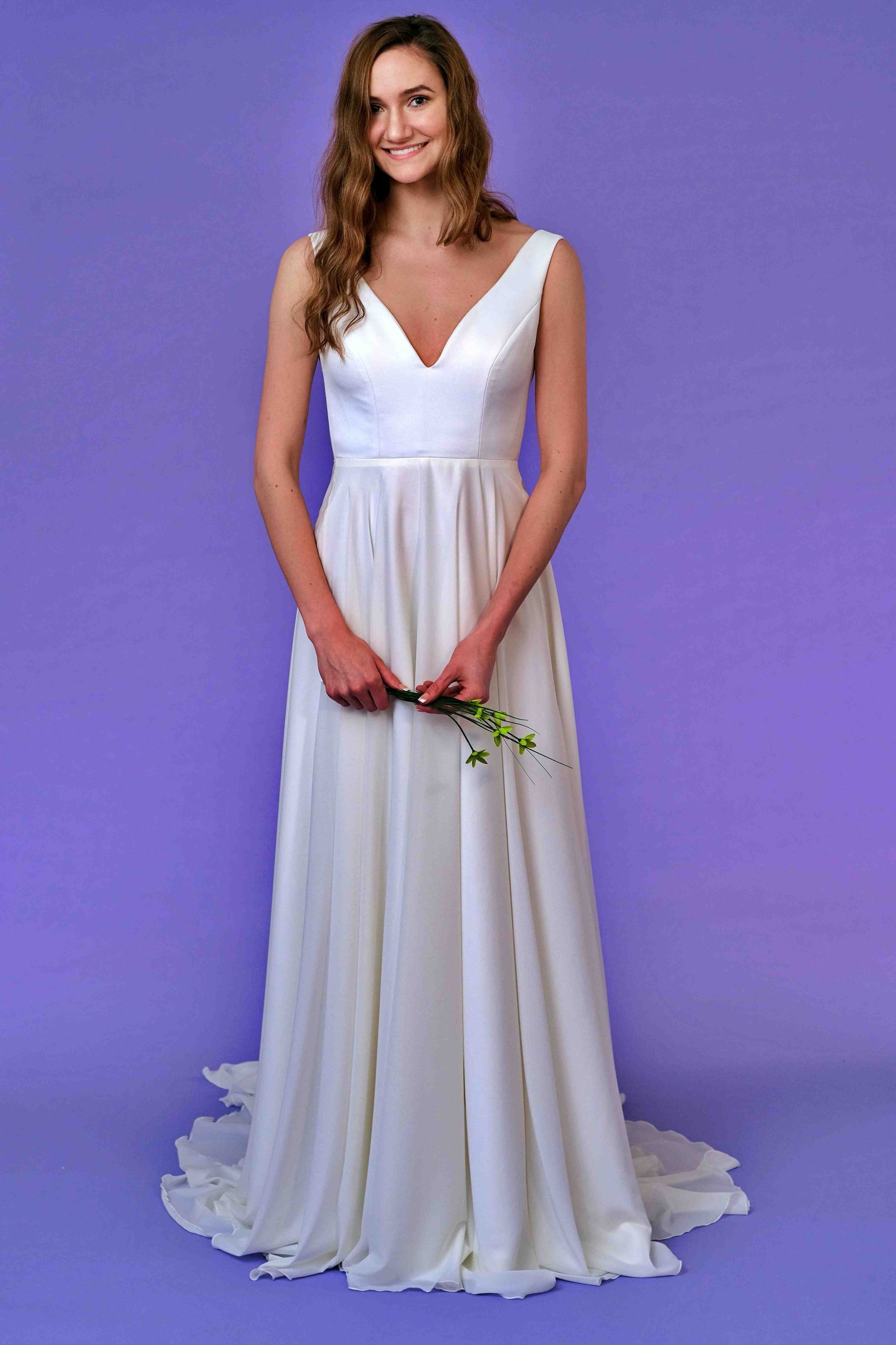 Woman in a white wedding dress with a V-neckline, holding flowers, against a purple backdrop.