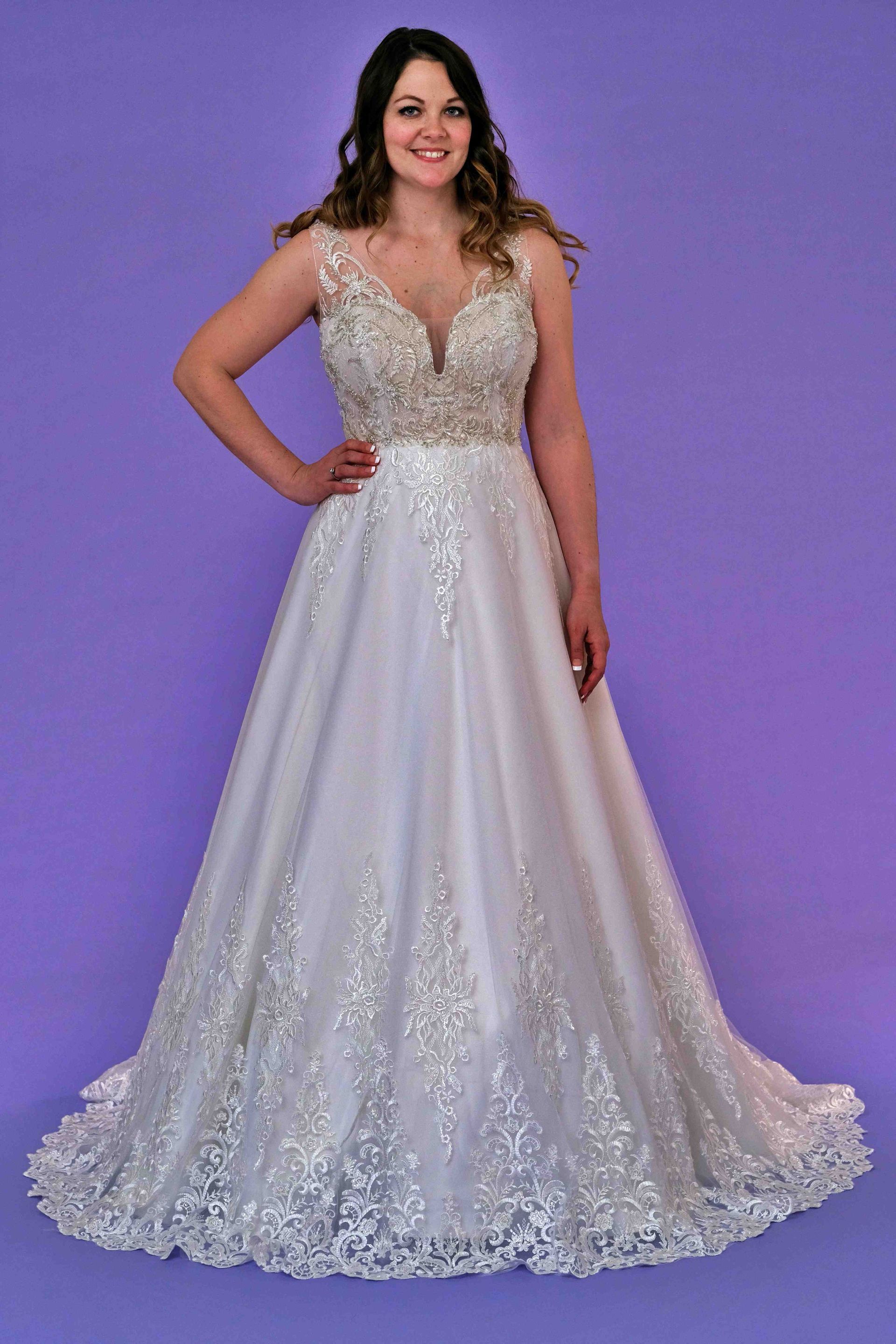 Woman in a white wedding dress with silver detailing and a long skirt, posing in front of a purple background.