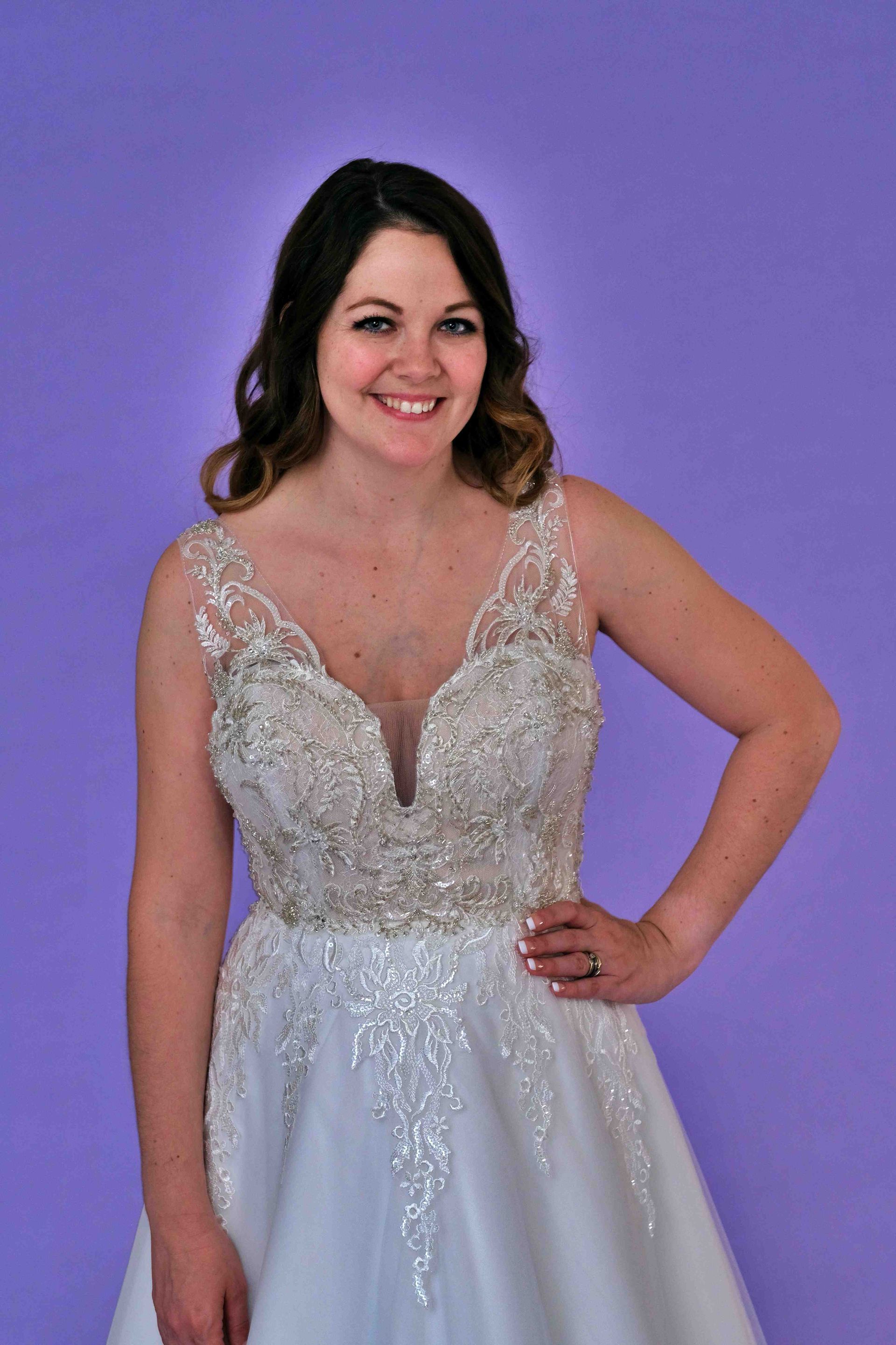 Woman in a white wedding dress with beading, smiling, posing against a purple backdrop.