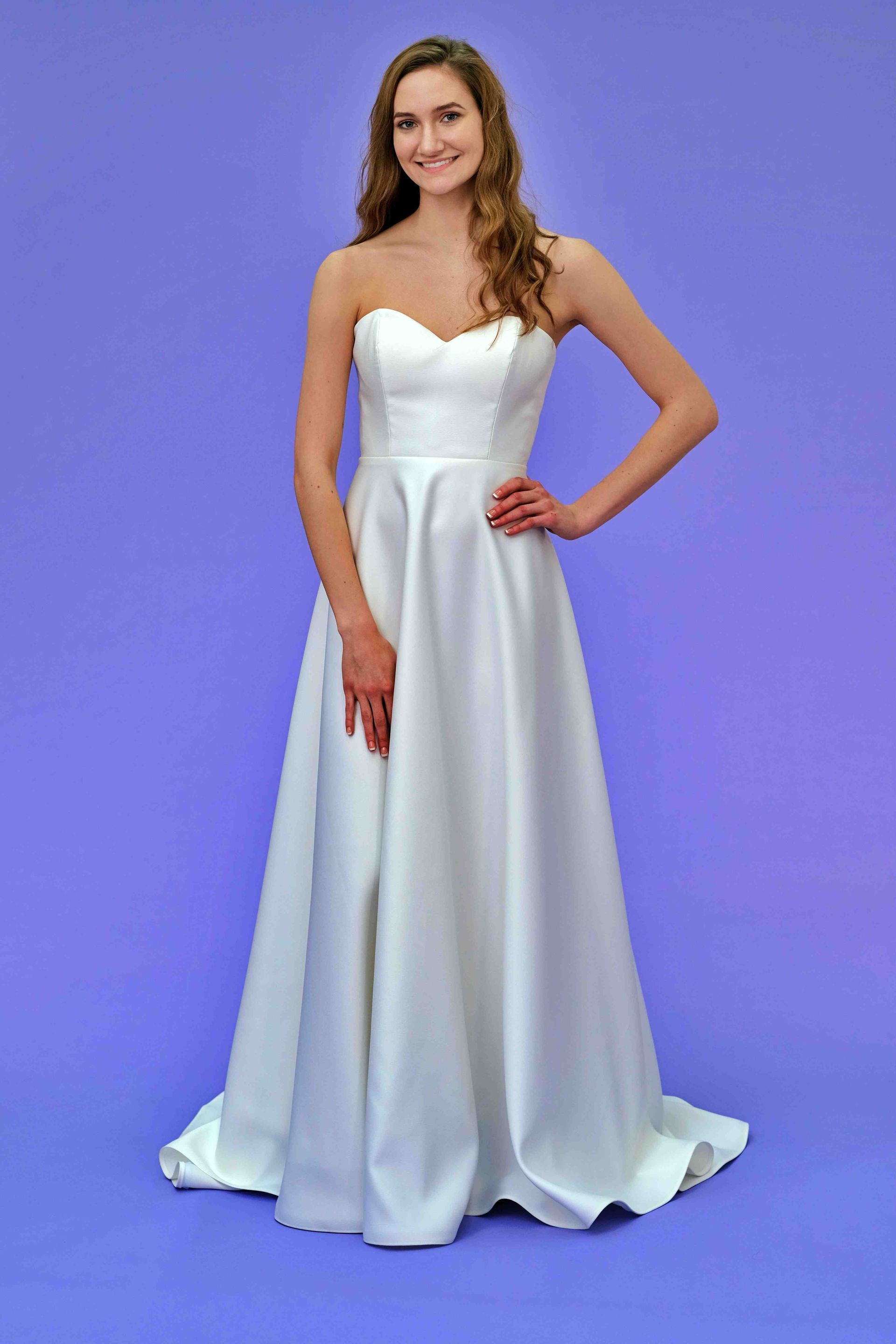 Woman in white strapless gown poses with hand on hip against a purple background.