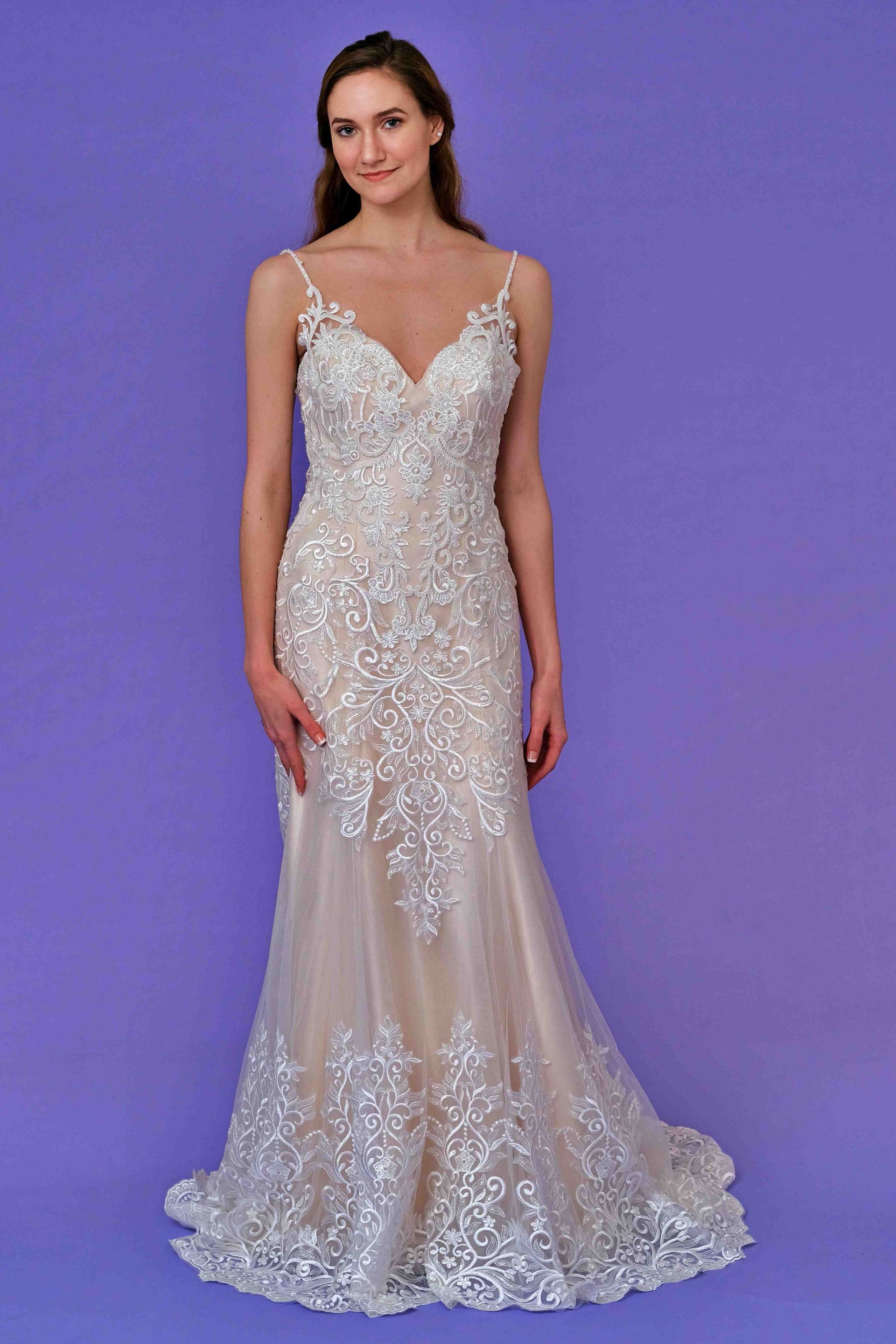 Woman in a beaded, ivory wedding dress with spaghetti straps and a mermaid silhouette, smiling in front of a purple backdrop.