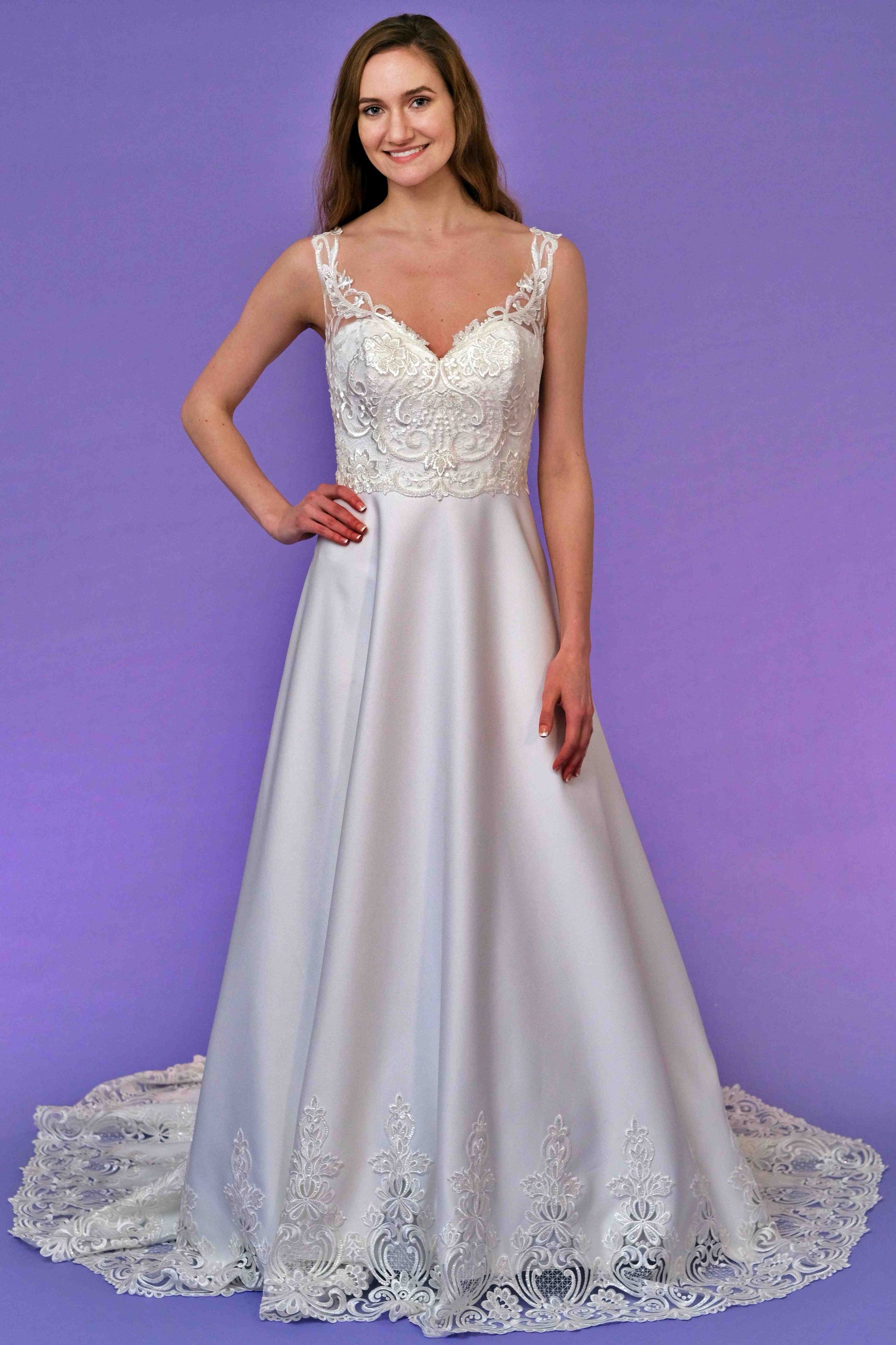 Woman in a white wedding dress with a beaded bodice and lace trim, posing in front of a purple background.