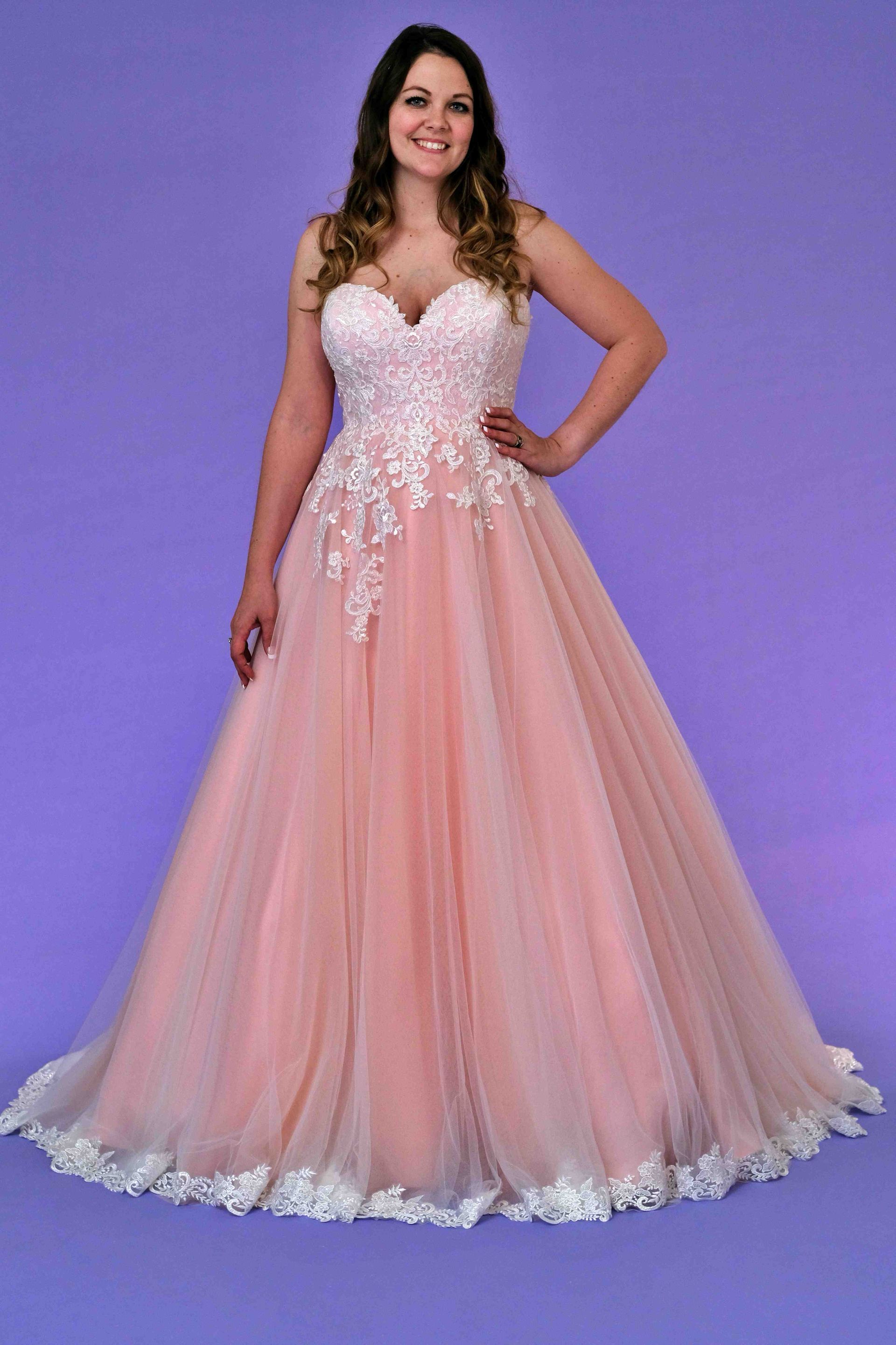 Woman in a pink and white ball gown, smiling, hand on hip, in front of a purple backdrop.