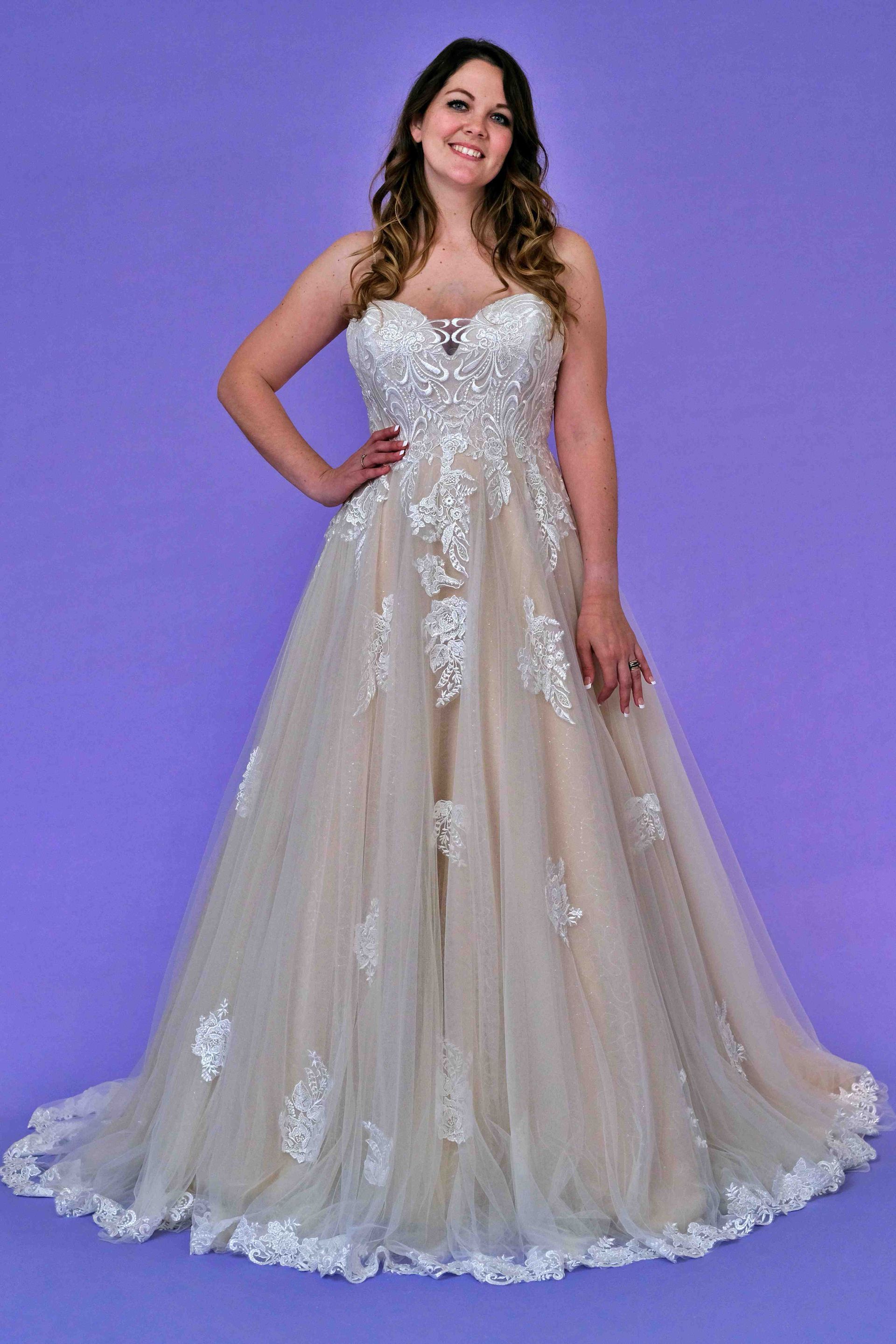 Woman in a strapless ivory and lace wedding gown, smiling, hand on hip, on a purple backdrop.