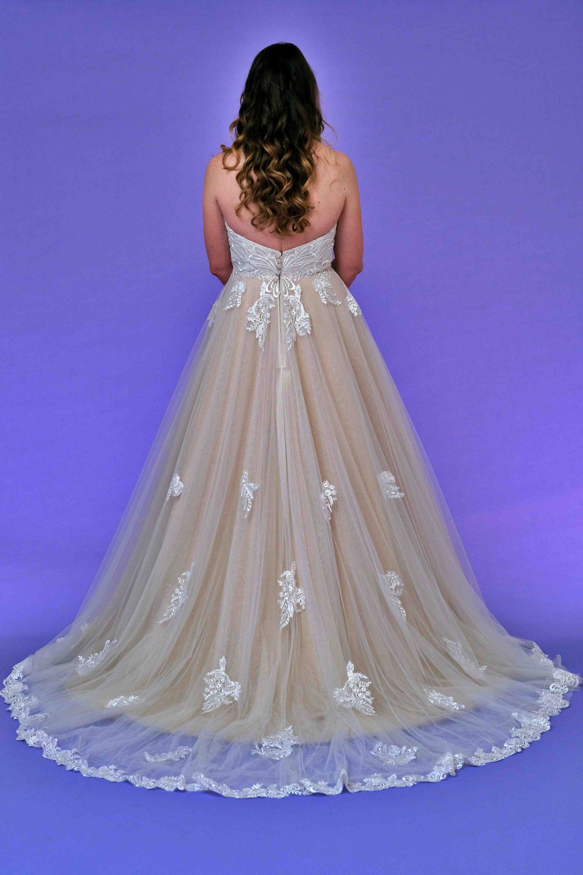 Woman in a strapless ivory and blush gown with a long train, standing against a purple background.