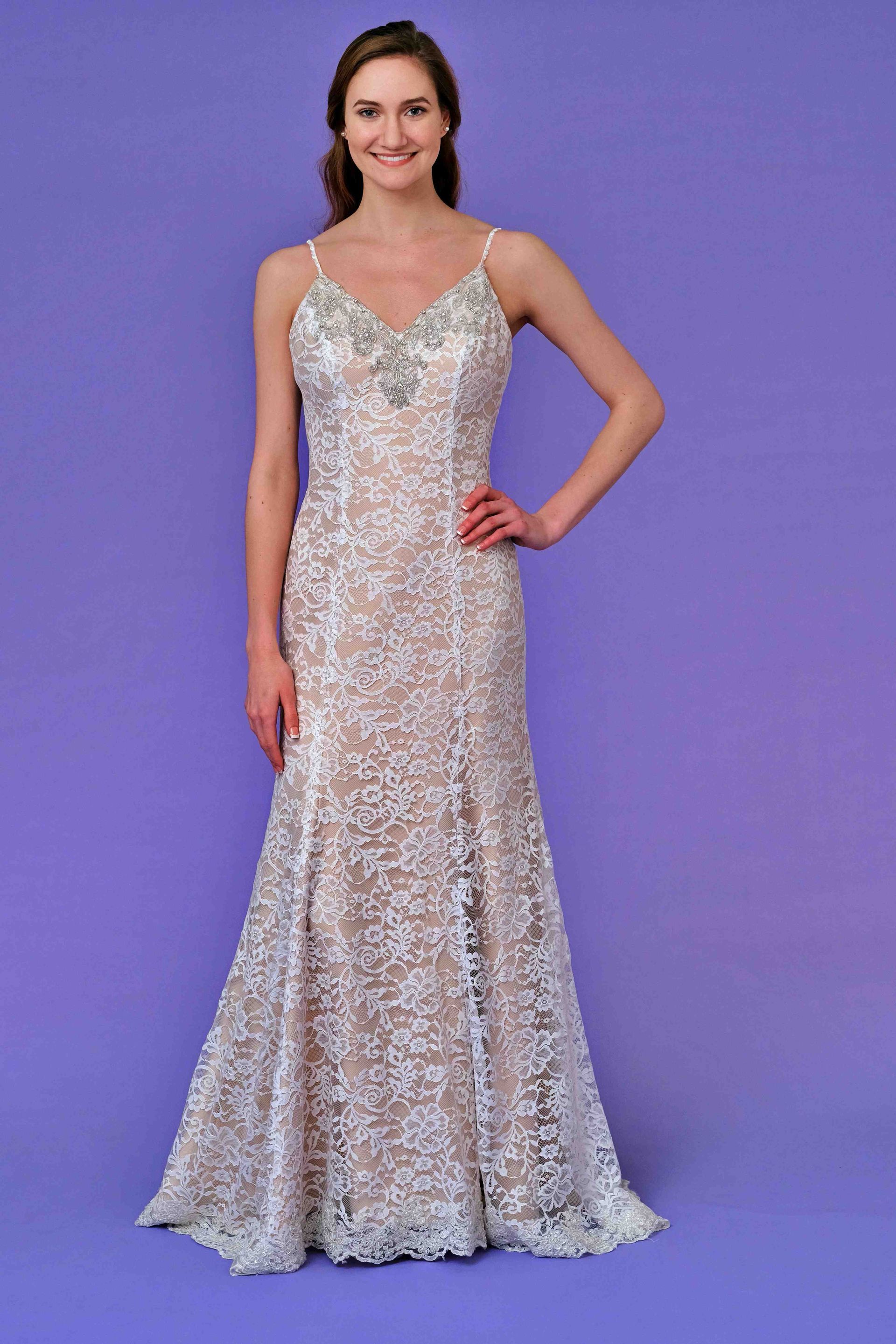 Woman in a white lace evening gown with beaded straps, standing against a purple background, hand on hip.