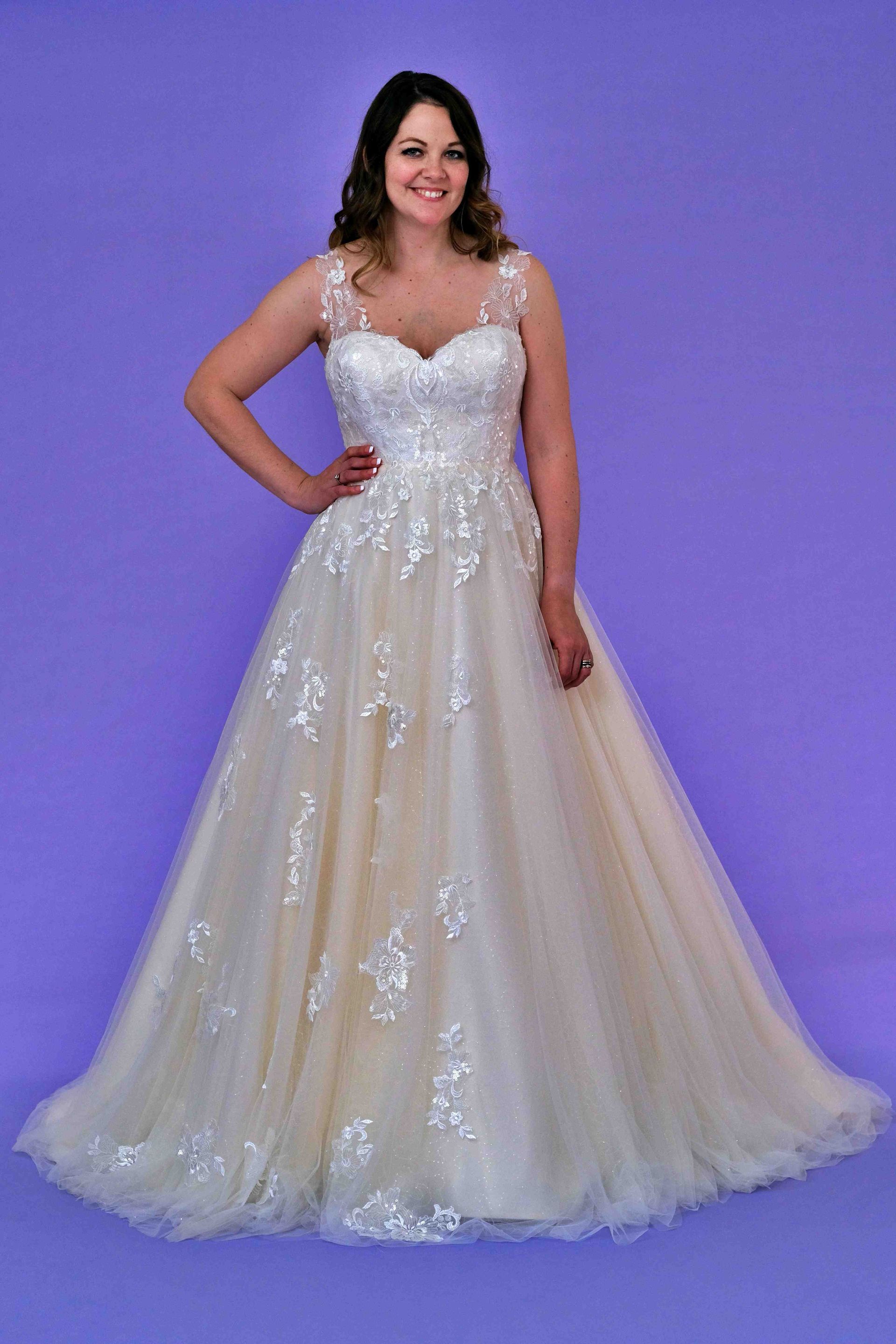 Woman in a white wedding gown, smiling, hand on hip, against a purple backdrop.