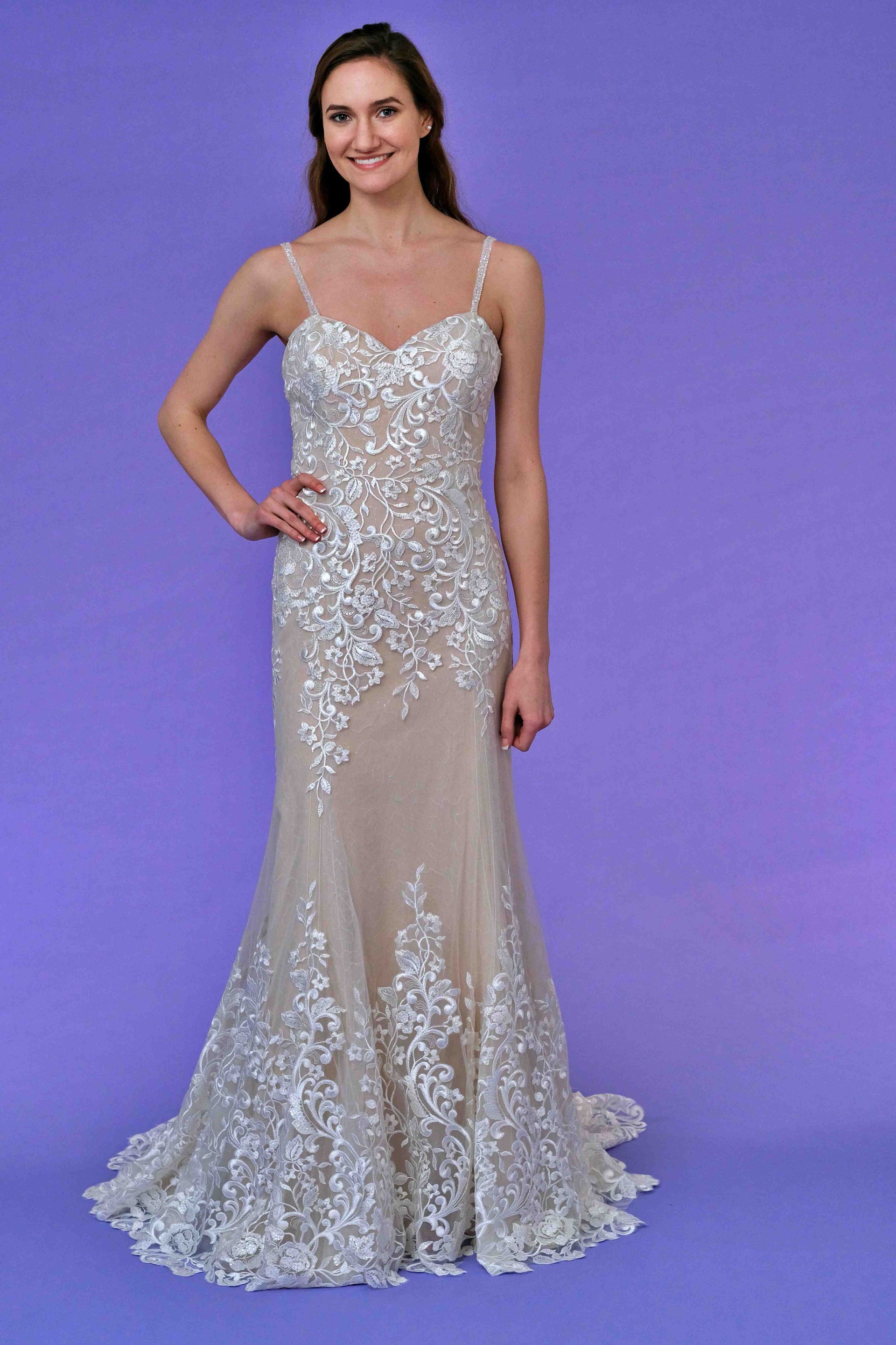 Woman in a wedding dress with beaded lace, thin straps, and a mermaid silhouette, standing in front of a purple backdrop.