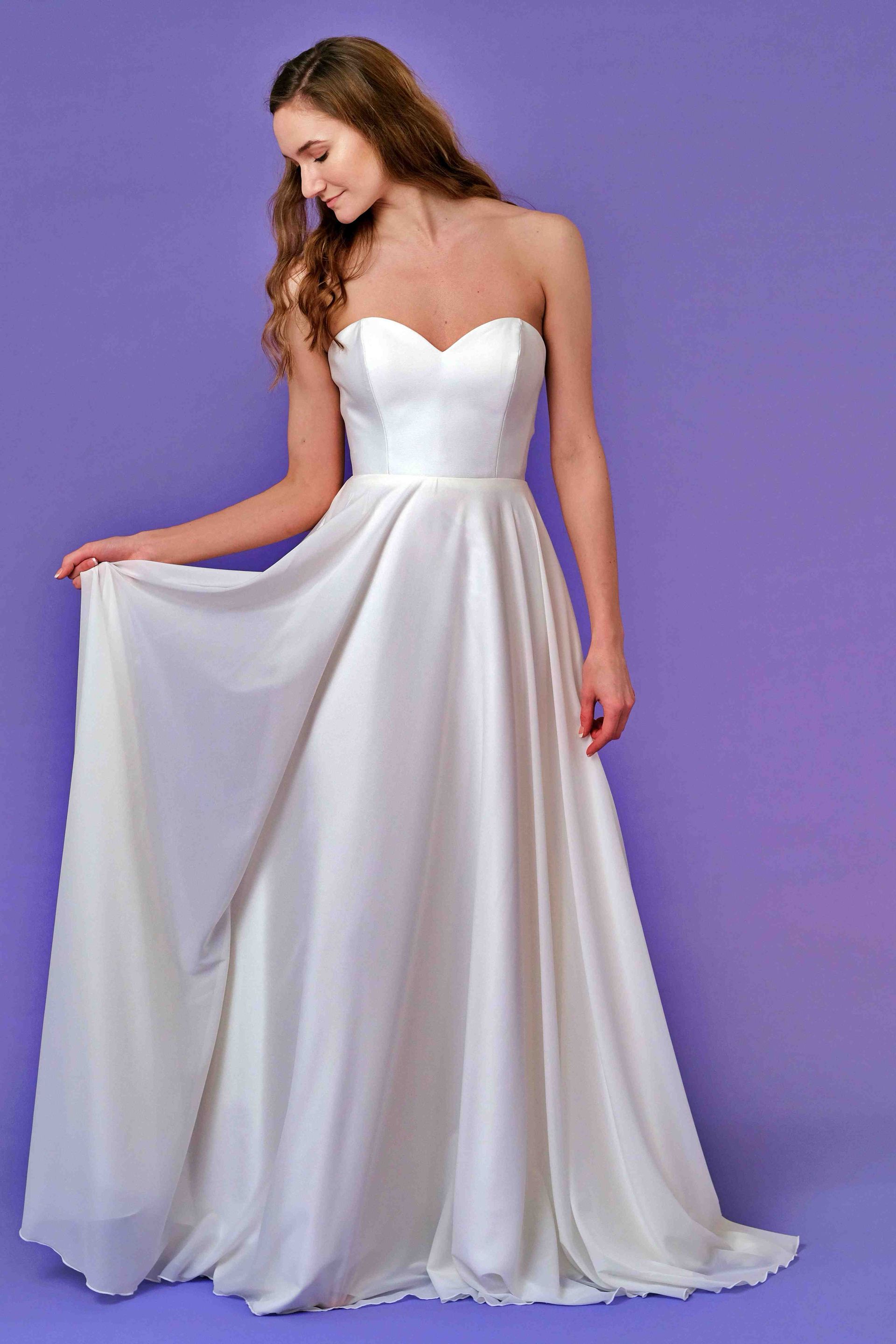 Woman in white strapless dress, holding up the skirt, on a purple backdrop.