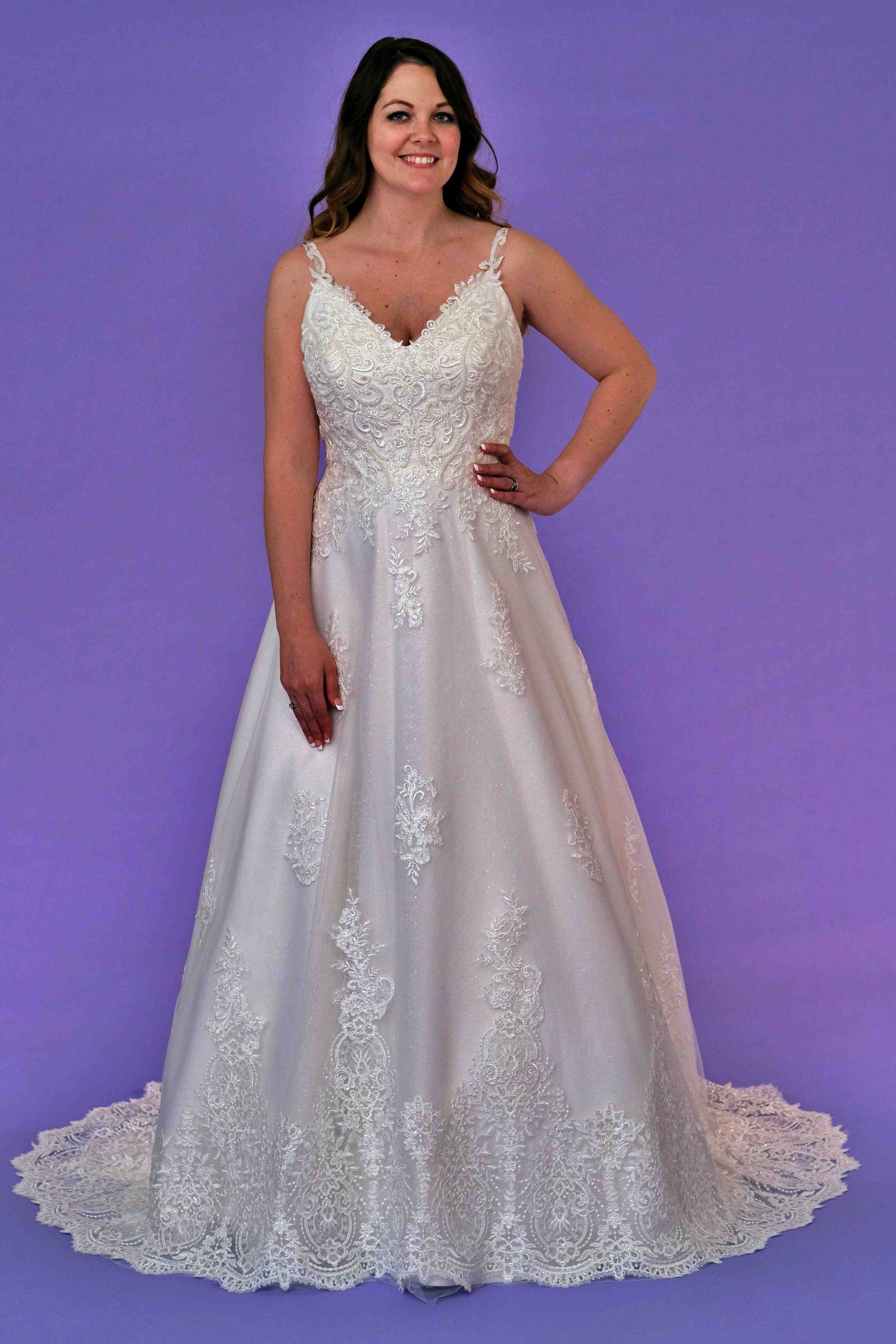 Woman in a white wedding dress with lace details, standing against a purple background, smiling.