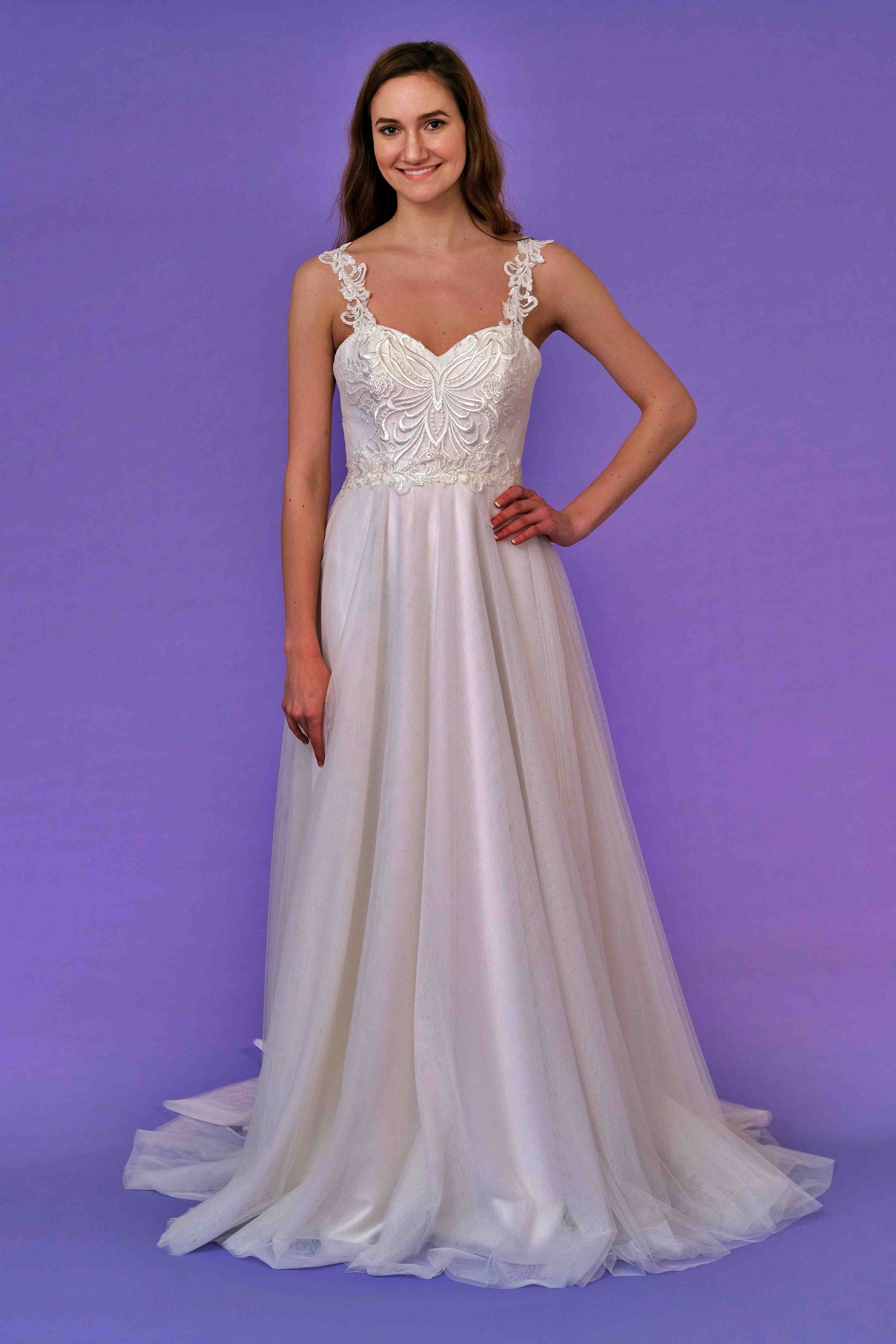 Woman in white wedding dress with floral bodice and thin straps, standing against a purple background.