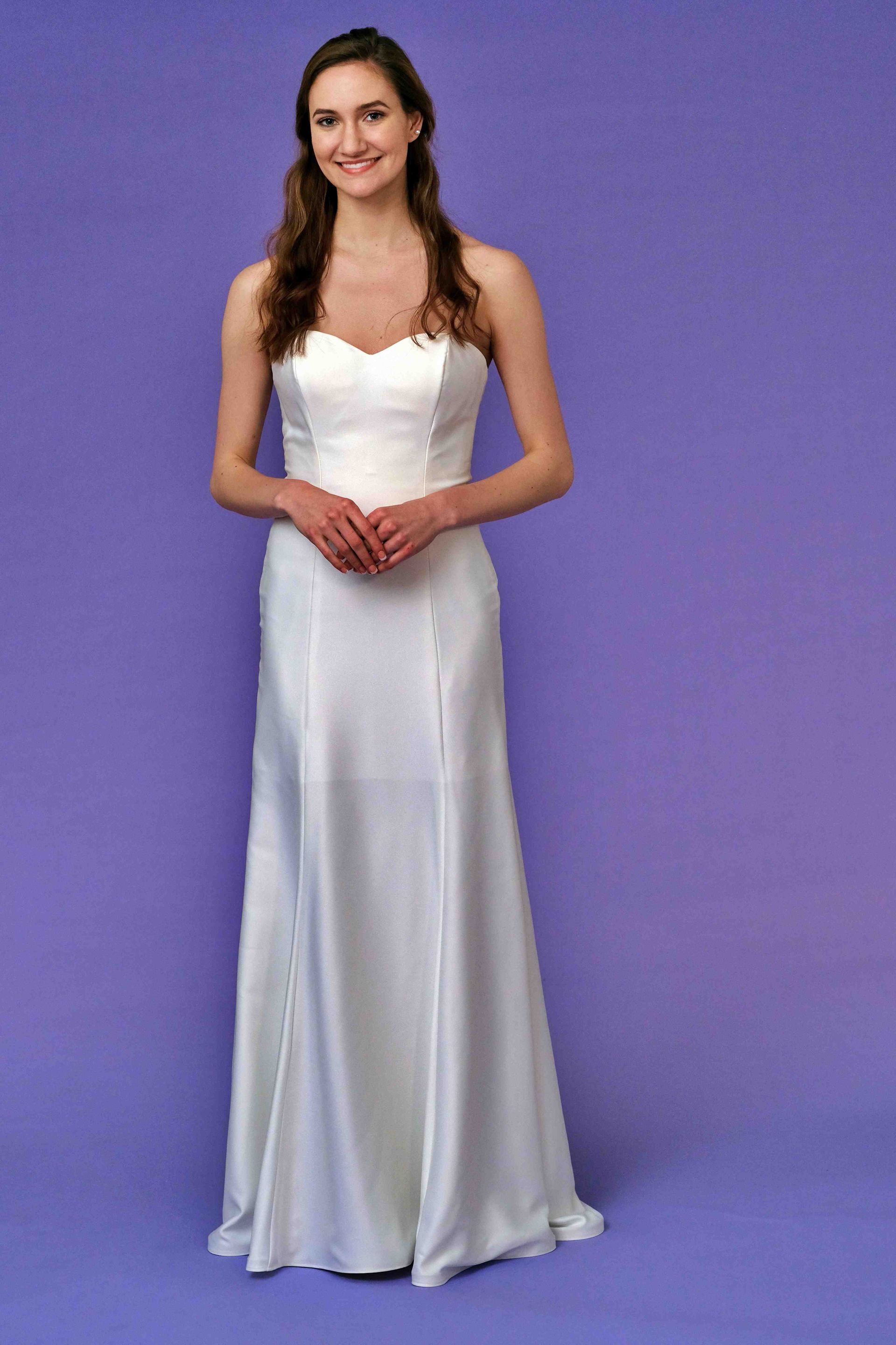 Woman in white strapless gown smiles against purple backdrop.