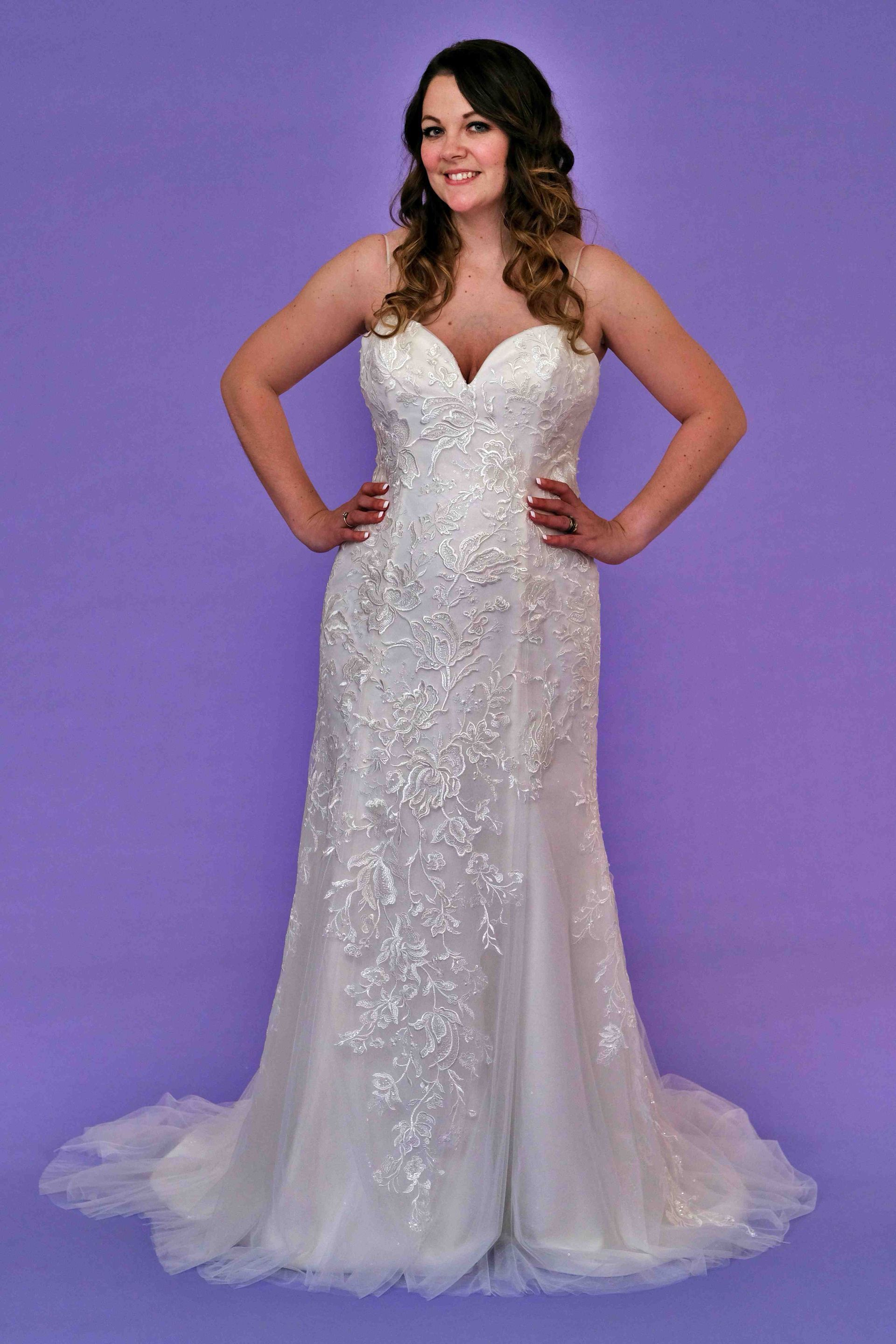 Woman in a white strapless wedding dress, hands on hips, smiling, purple background.