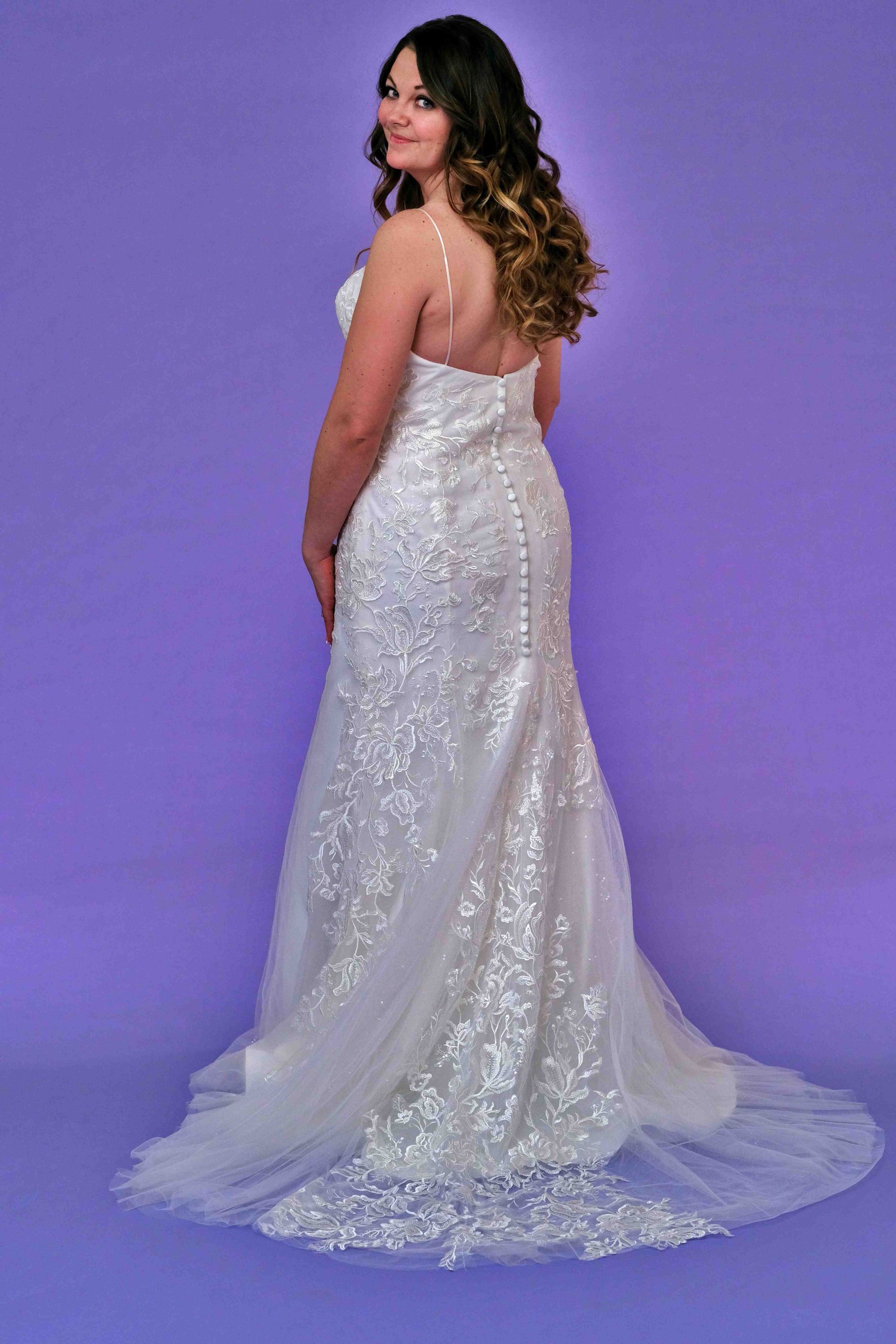 Woman in a white wedding dress with a train, standing against a purple backdrop, looking over her shoulder.