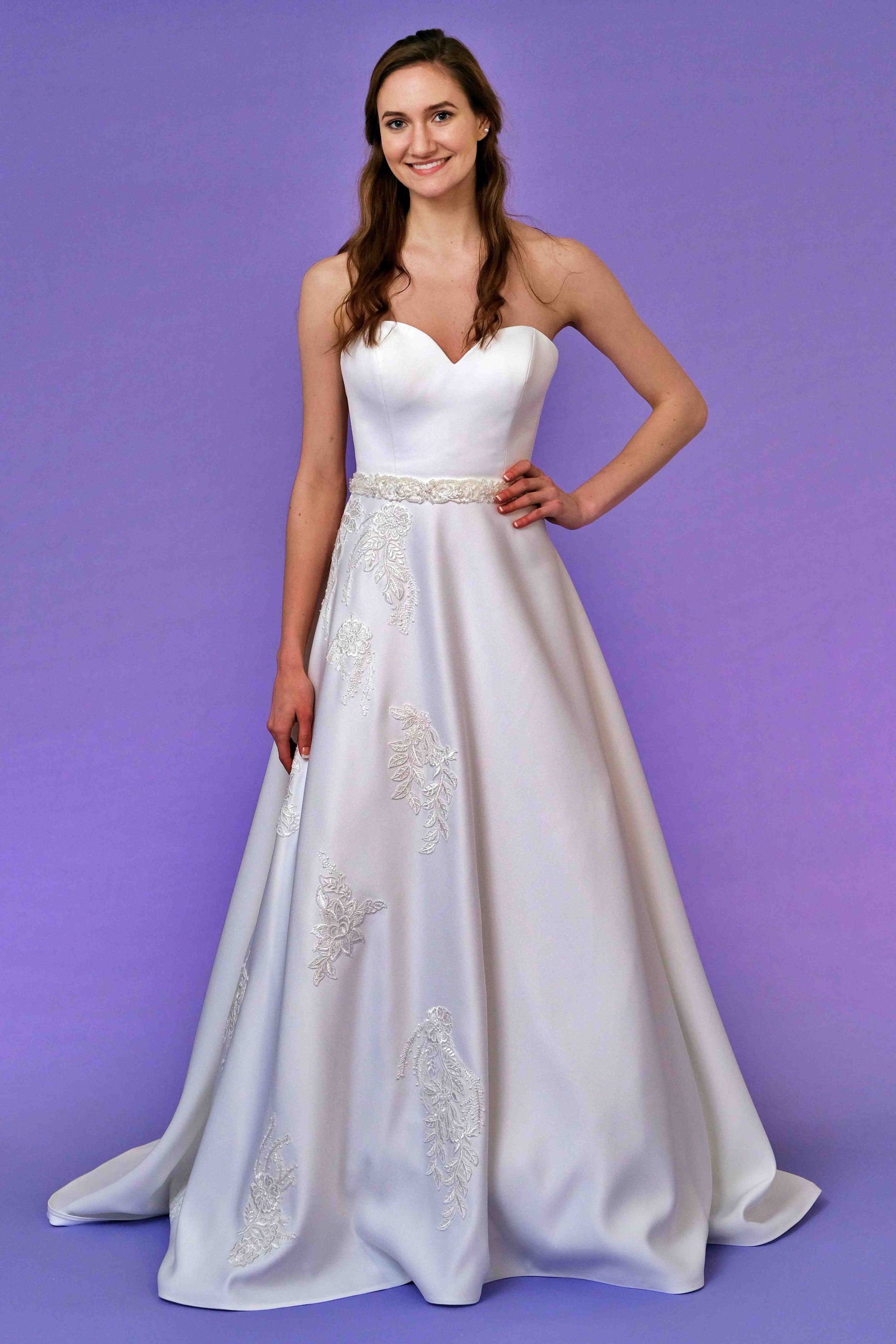 Woman in strapless white wedding dress with beaded accents, standing in front of a purple backdrop, smiling.