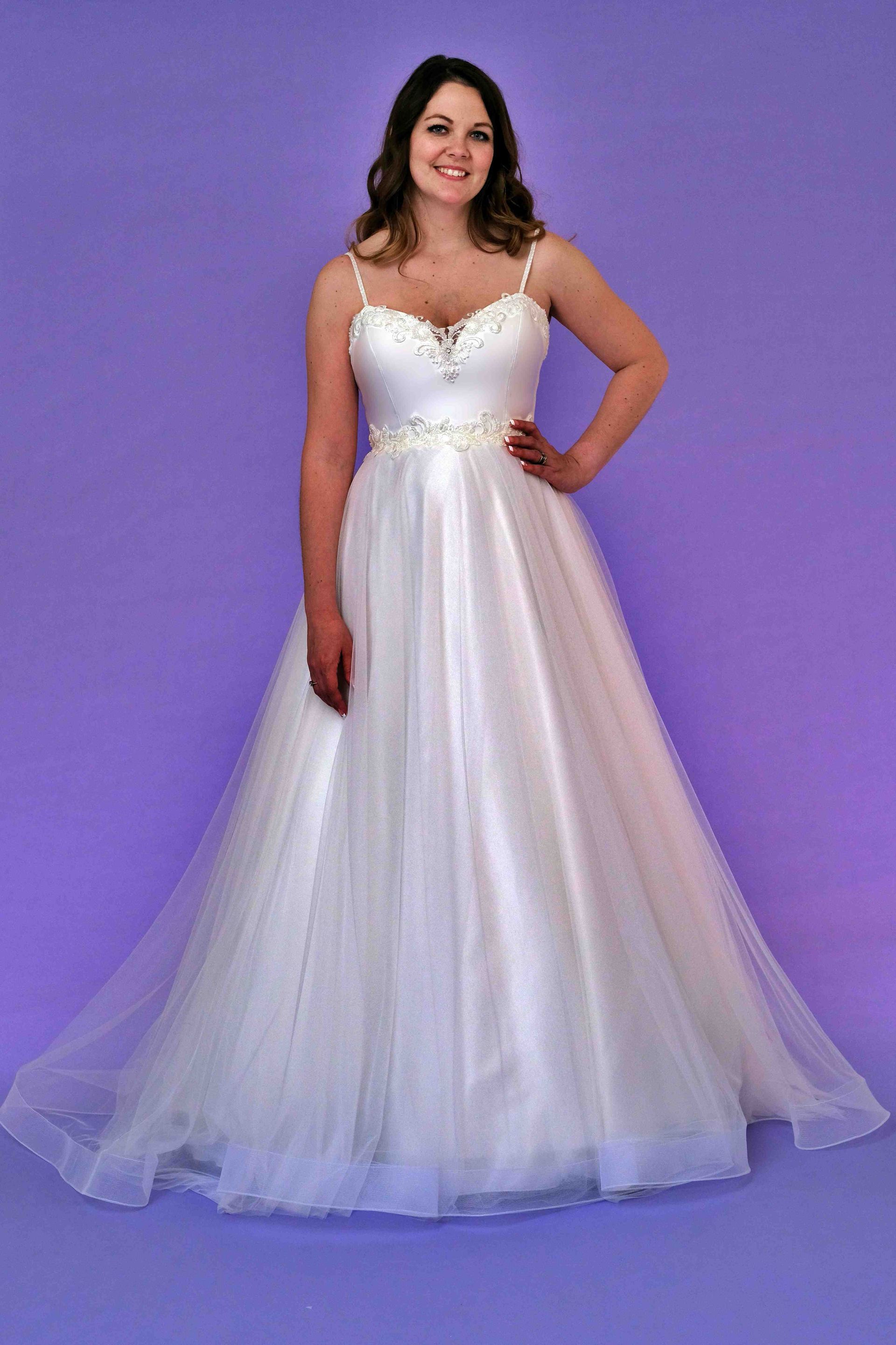 Woman wearing a white ball gown with a beaded bodice and spaghetti straps, posing in front of a purple backdrop.