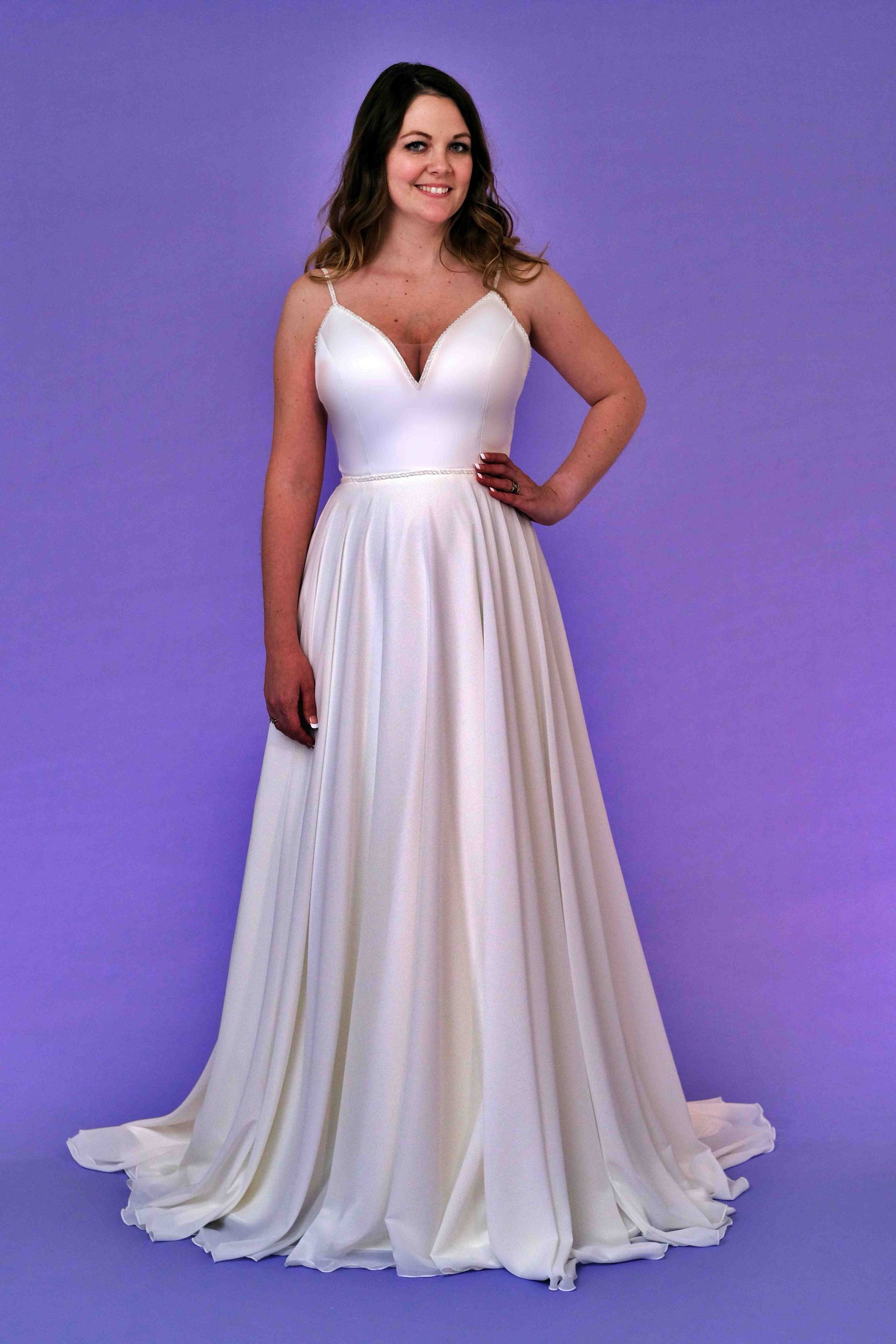 Woman in a white wedding dress with a low neckline, posing with hand on hip against a purple backdrop.