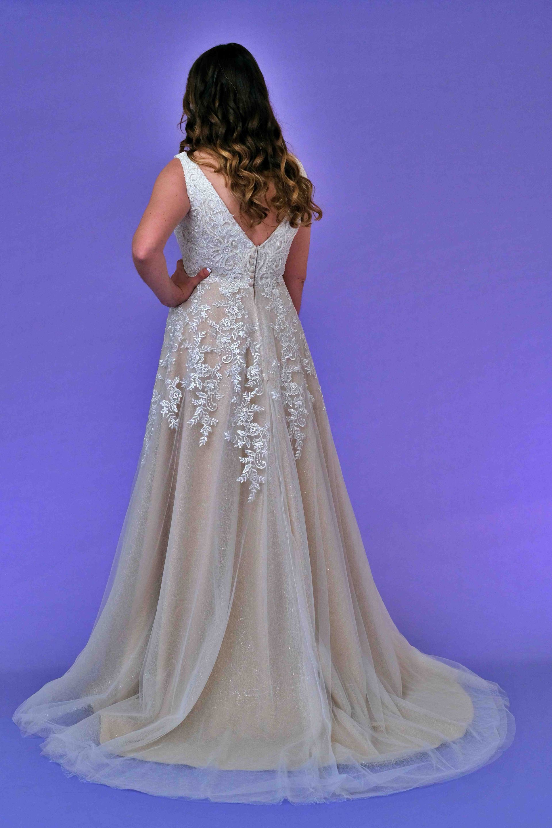 Woman in a beaded, light-colored gown poses against a purple background, hand on hip, back to camera.