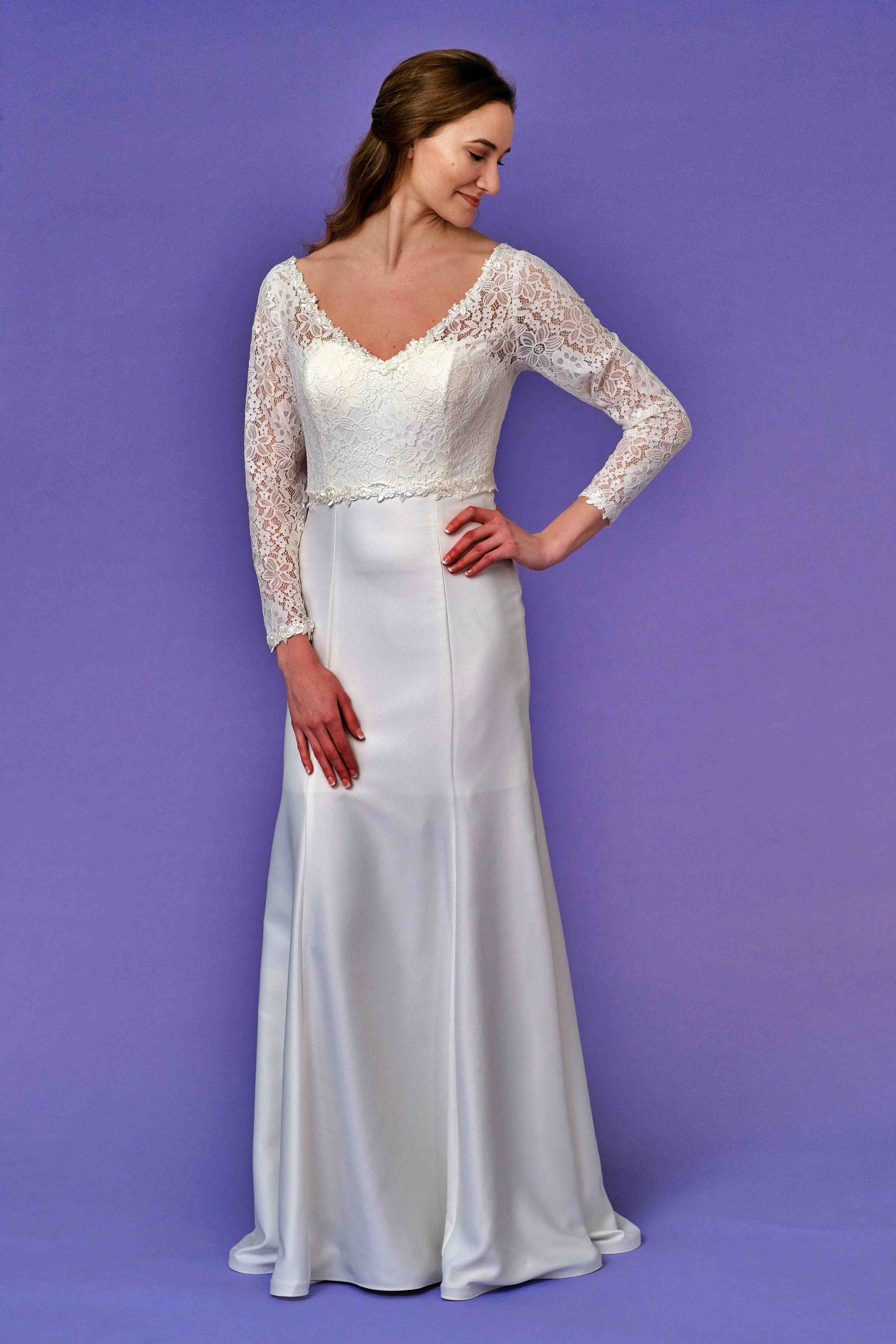 Woman in white wedding dress with lace bodice, hand on hip, smiling, purple background.