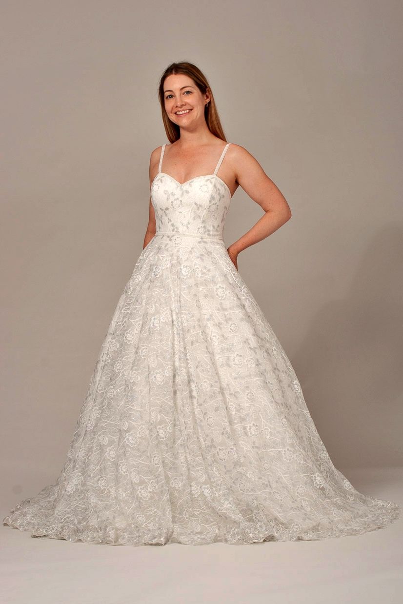 Woman in a white wedding gown, smiling, standing in front of a plain background.