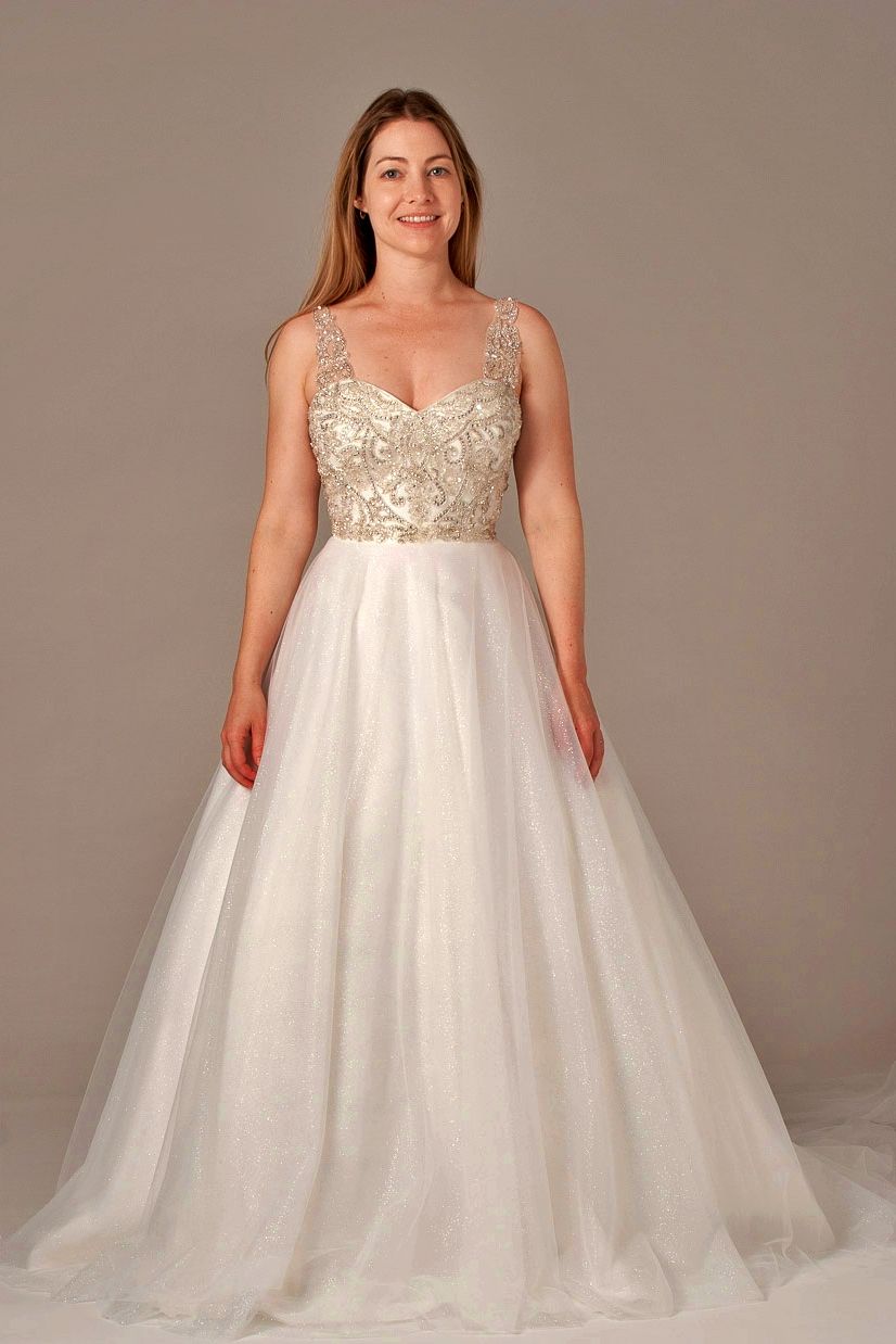 Woman in a white wedding gown with a beaded bodice, standing in front of a neutral backdrop.