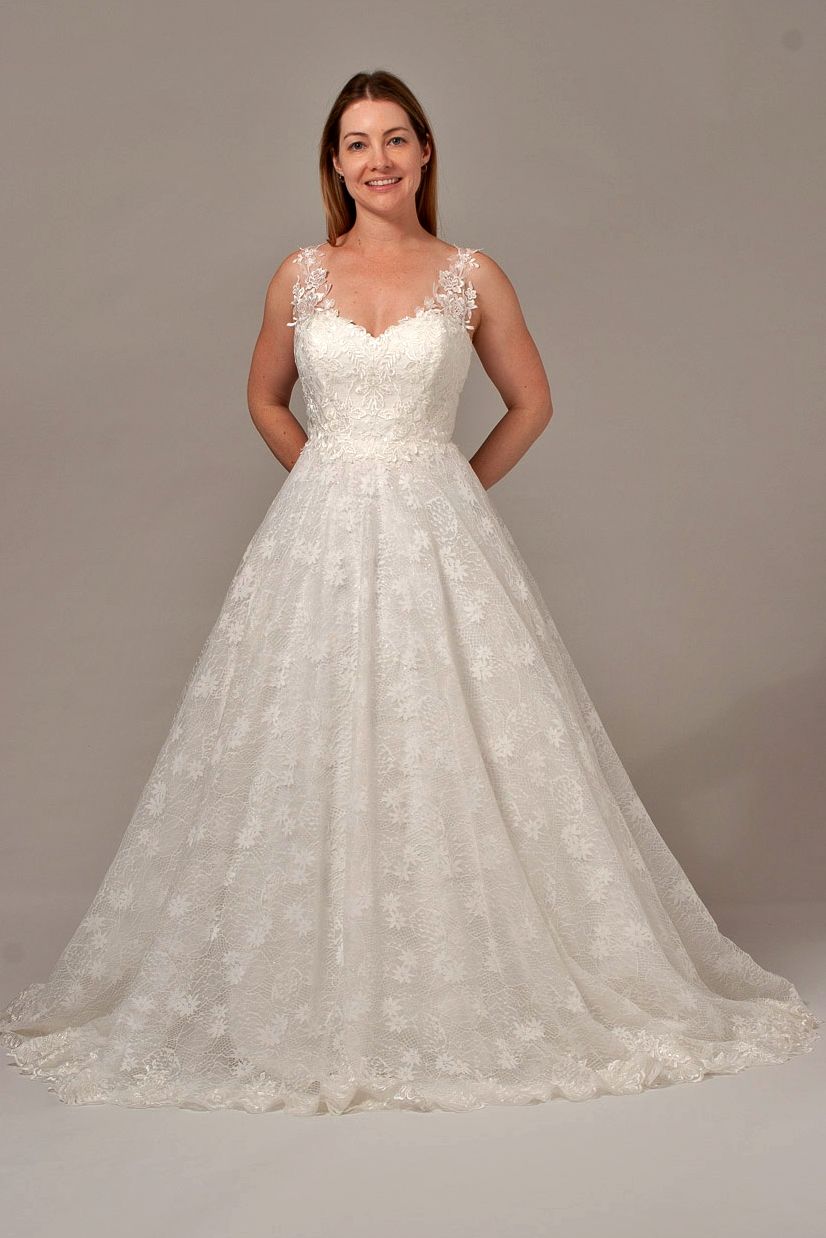Woman in white lace wedding dress, hands behind back, smiling.
