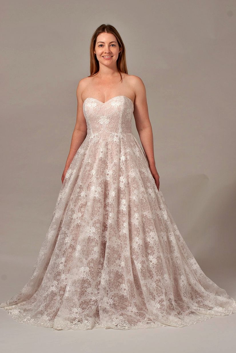 Woman wearing a strapless, floral-lace wedding dress, standing indoors, smiling.