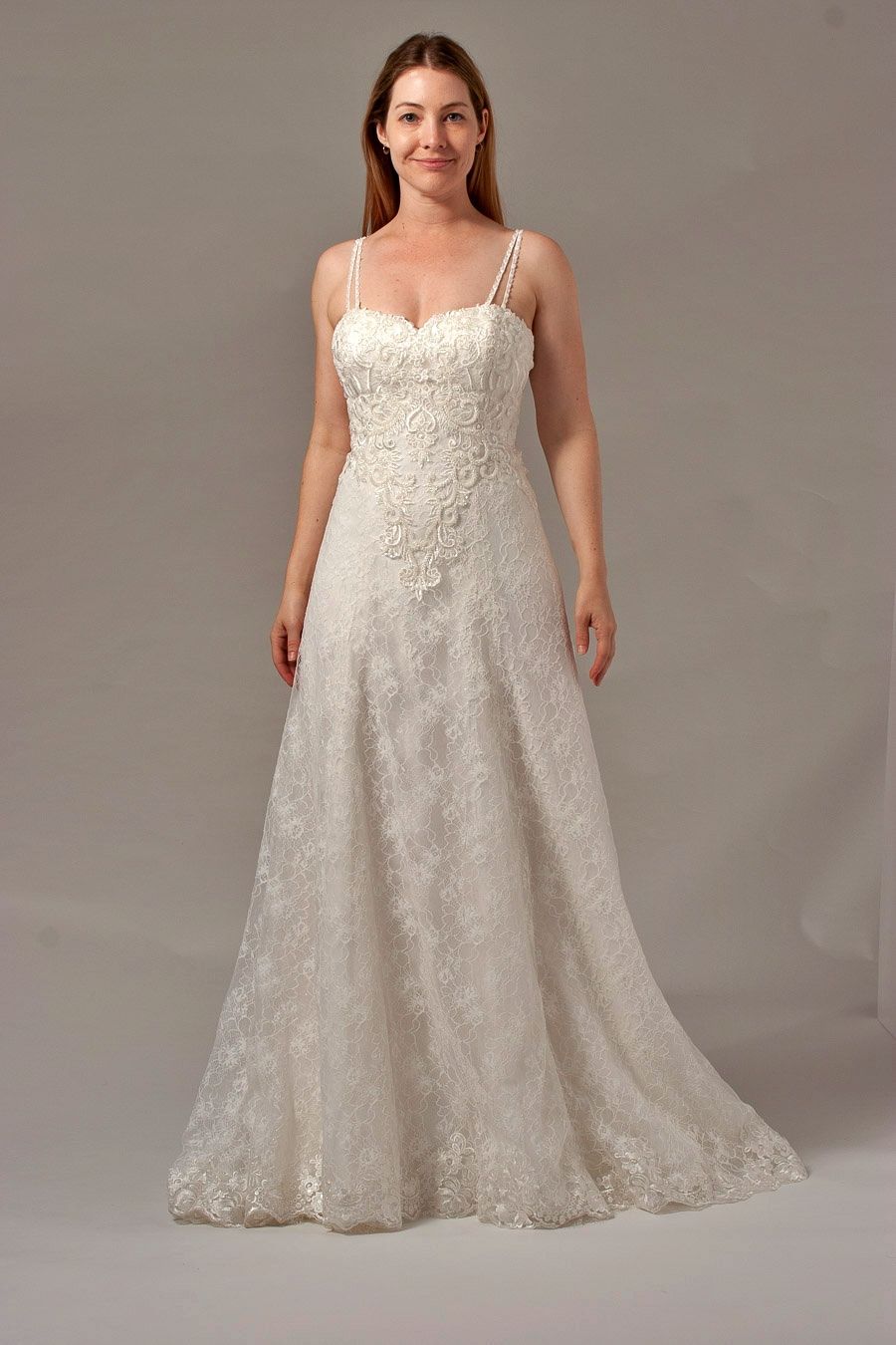 Woman in a white wedding dress with a beaded bodice and spaghetti straps, standing against a neutral background.